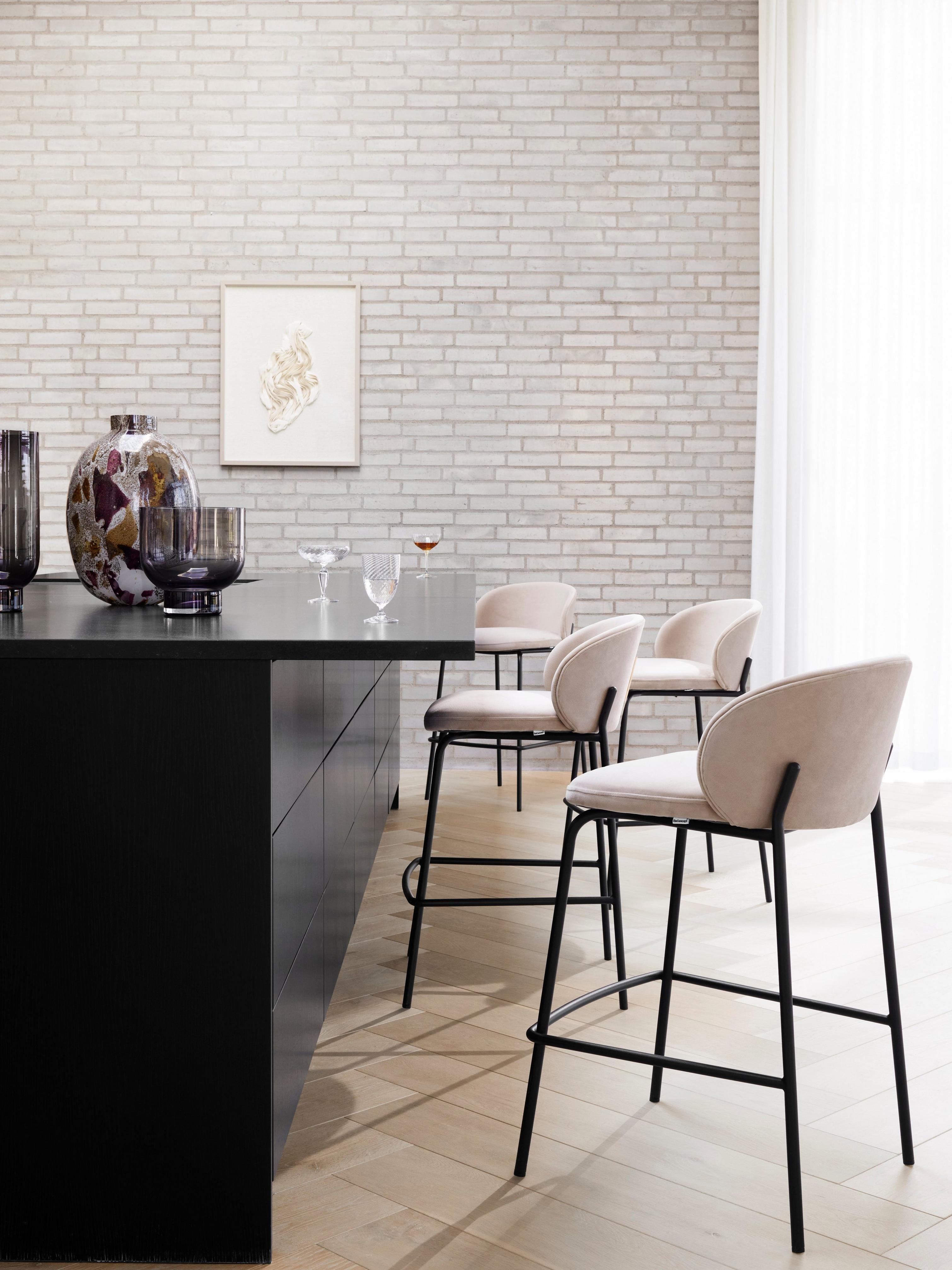Modern kitchen bar with black countertop, beige high chairs, and decorative vases against a white brick wall.