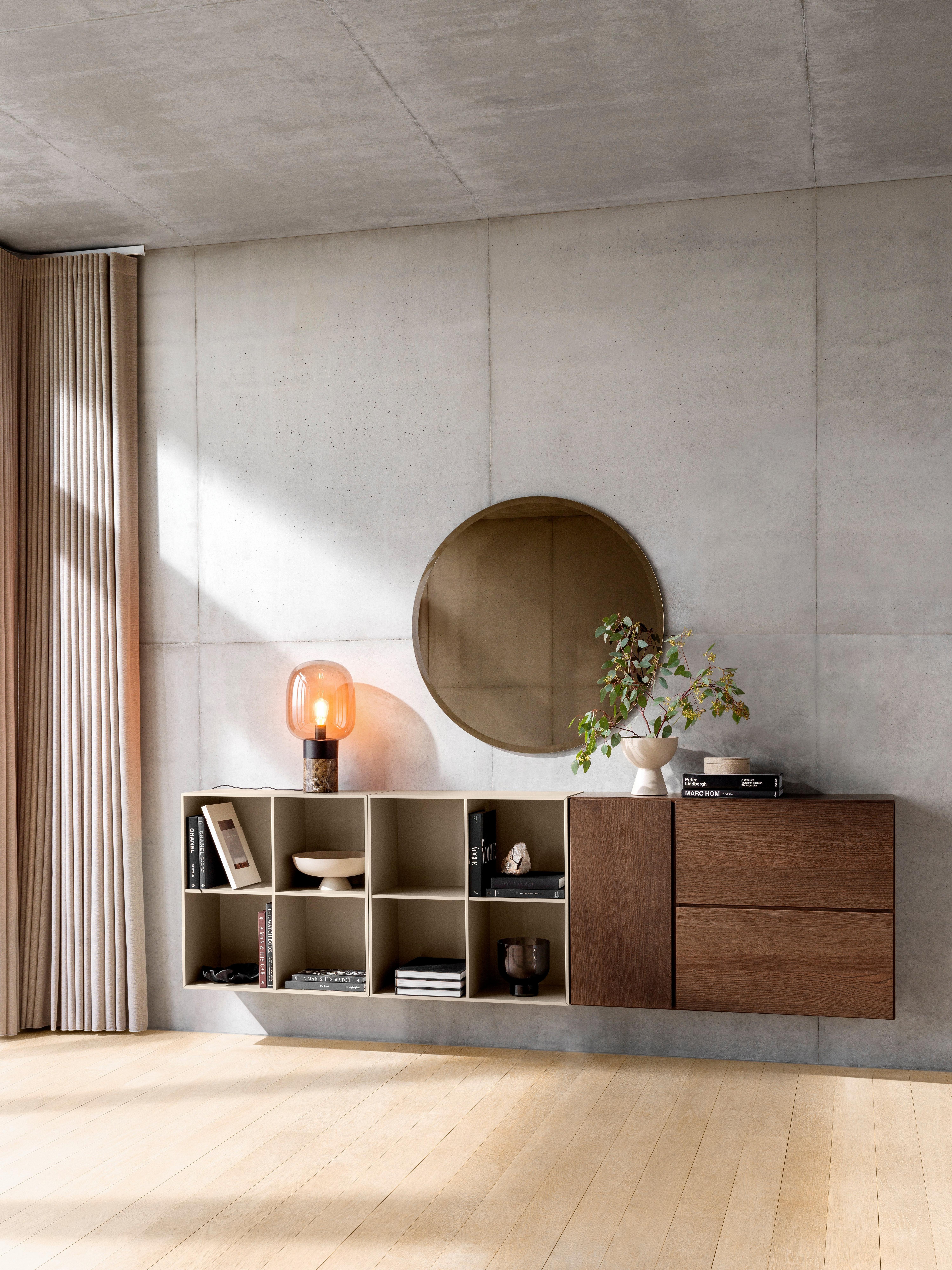 Wall-mounted storage with beige open shelves and wood drawers, styled with books, decor, a round mirror, and a warm glass lamp