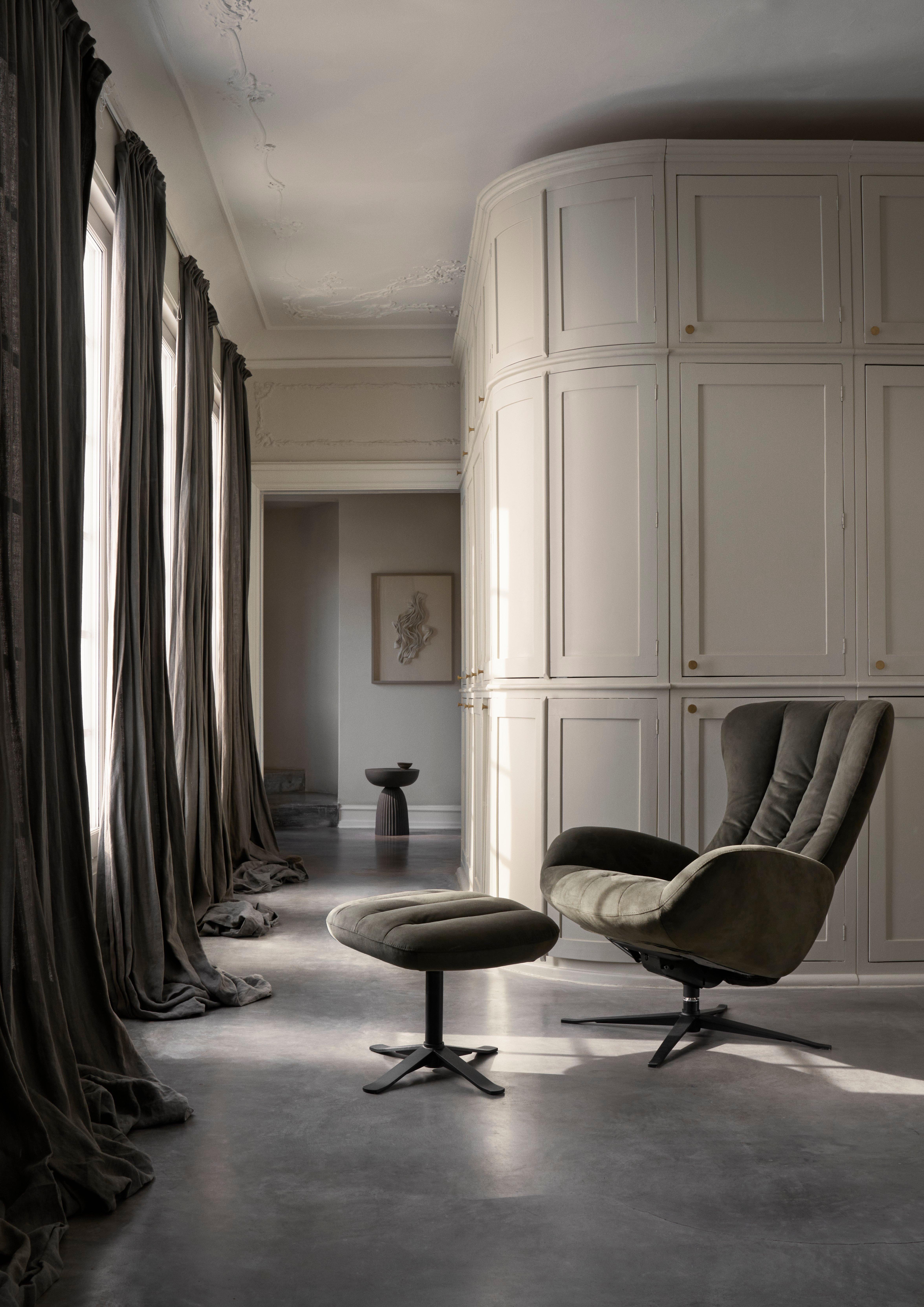 Dark green swivel armchair with matching ottoman on black bases, placed on grey concrete floor near tall windows with heavy curtains.