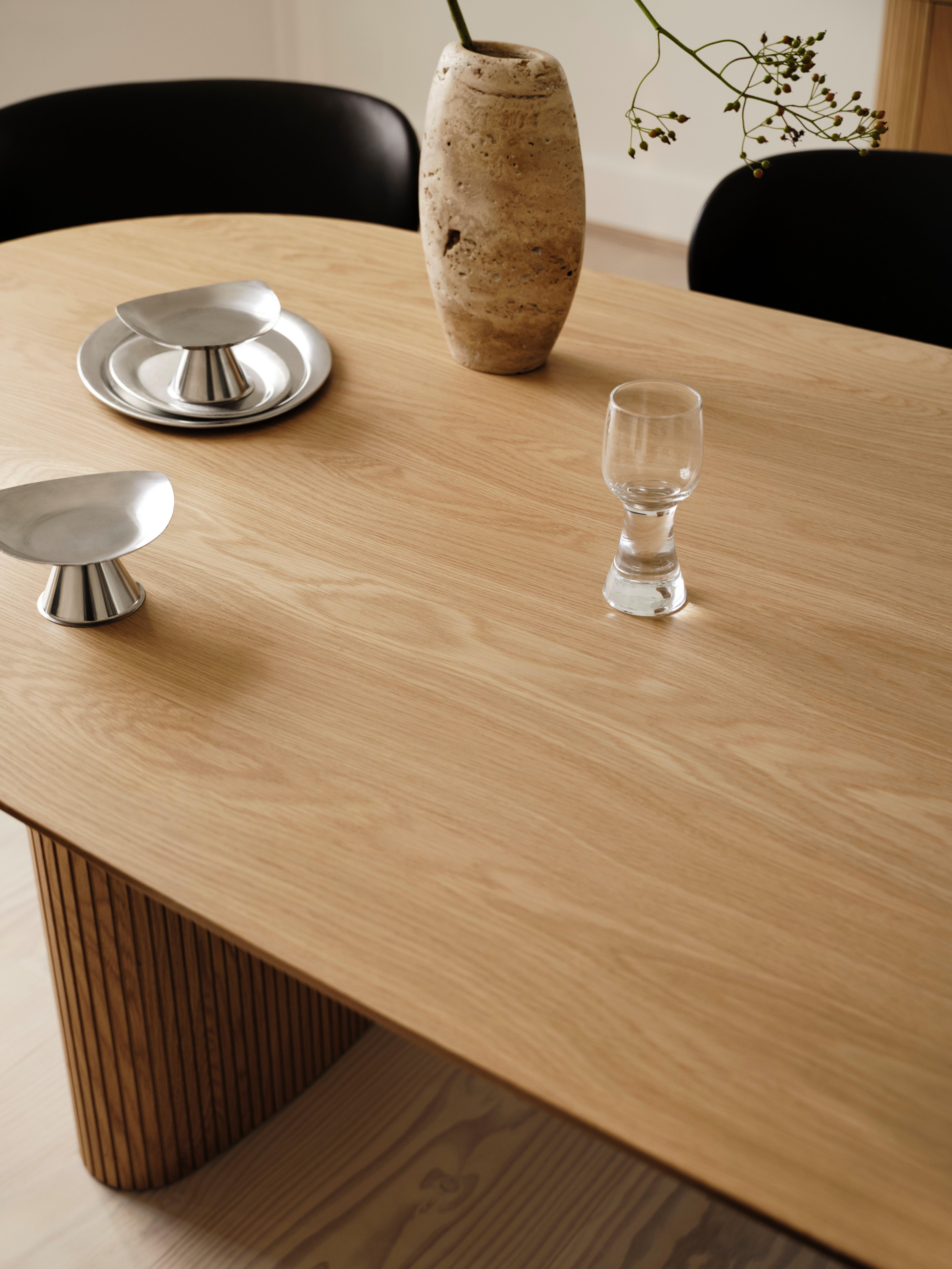 Wooden table with metal dishes, glass, and a stone vase holding a plant. Black chairs visible.
