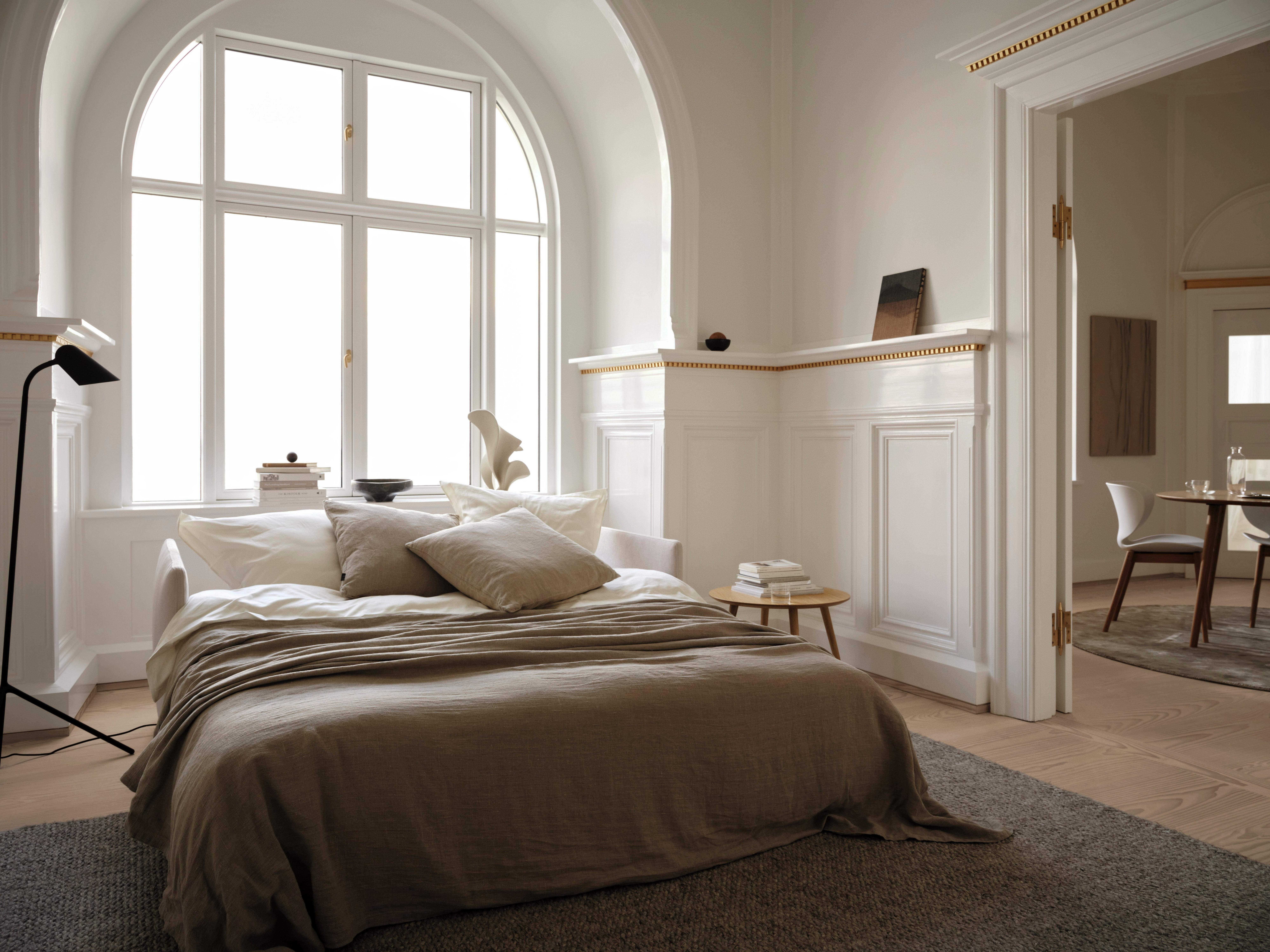 Cozy bedroom with a large window, bed, pillows, and a side table with books. Dining area visible.