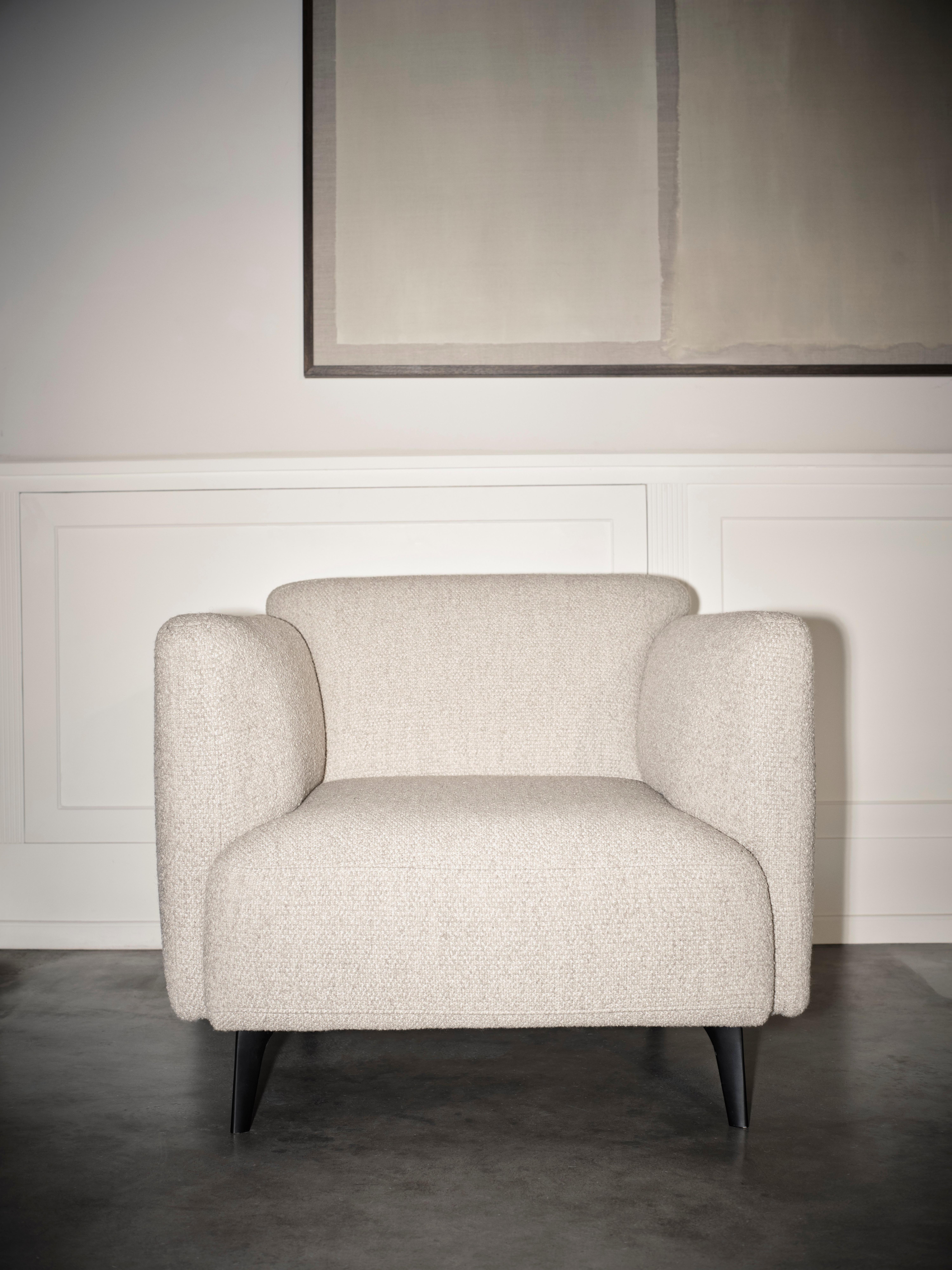 Contemporary armchair in textured light beige fabric with rounded arms and black tapered legs, set against a minimal wall panel.