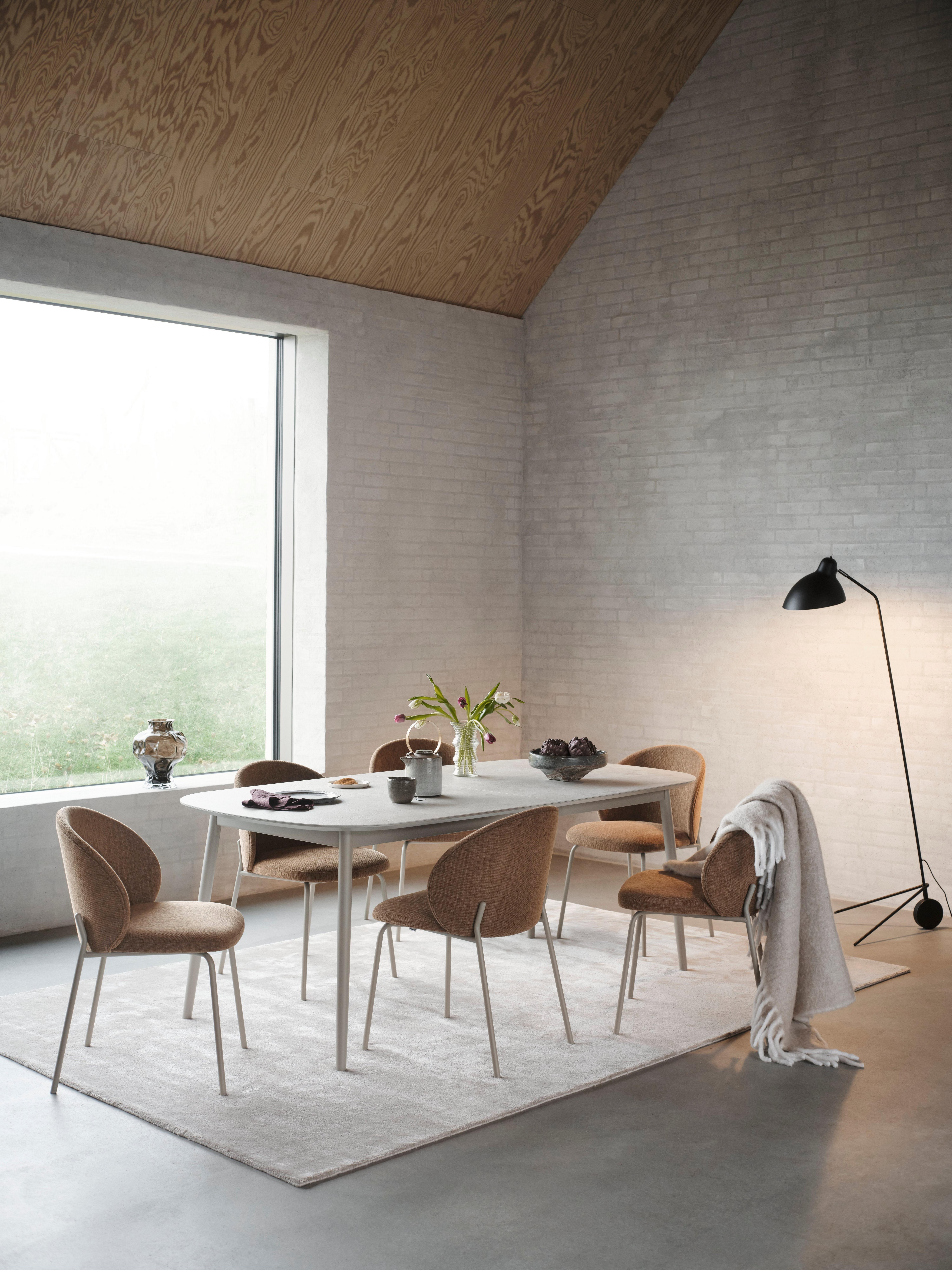 Dining area with six tan fabric chairs around a white table on a light rug, next to a large window and black floor lamp.
