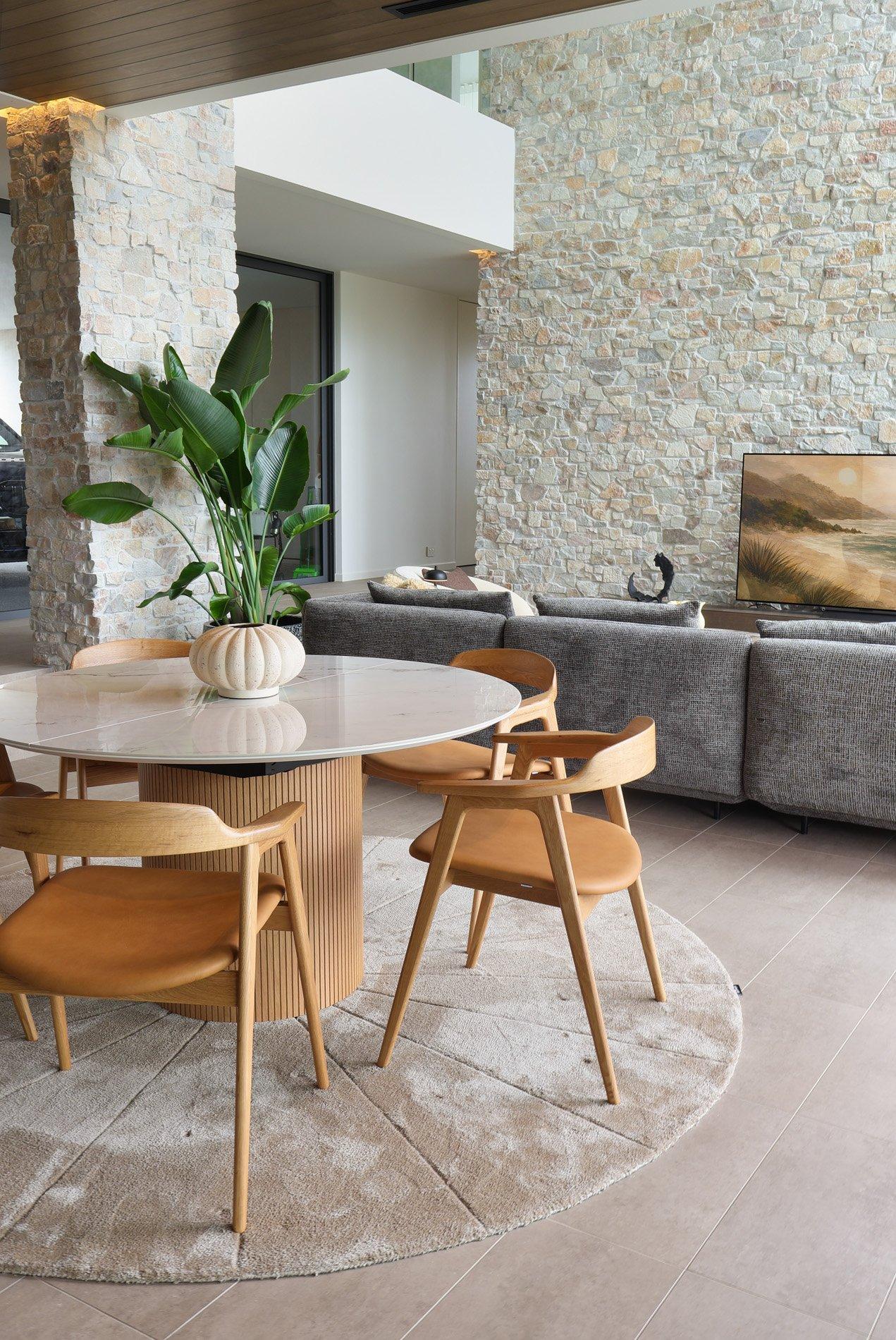 Rundt spisebord med trestoler, plante i vase, grå sofa og stort maleri på vegg.