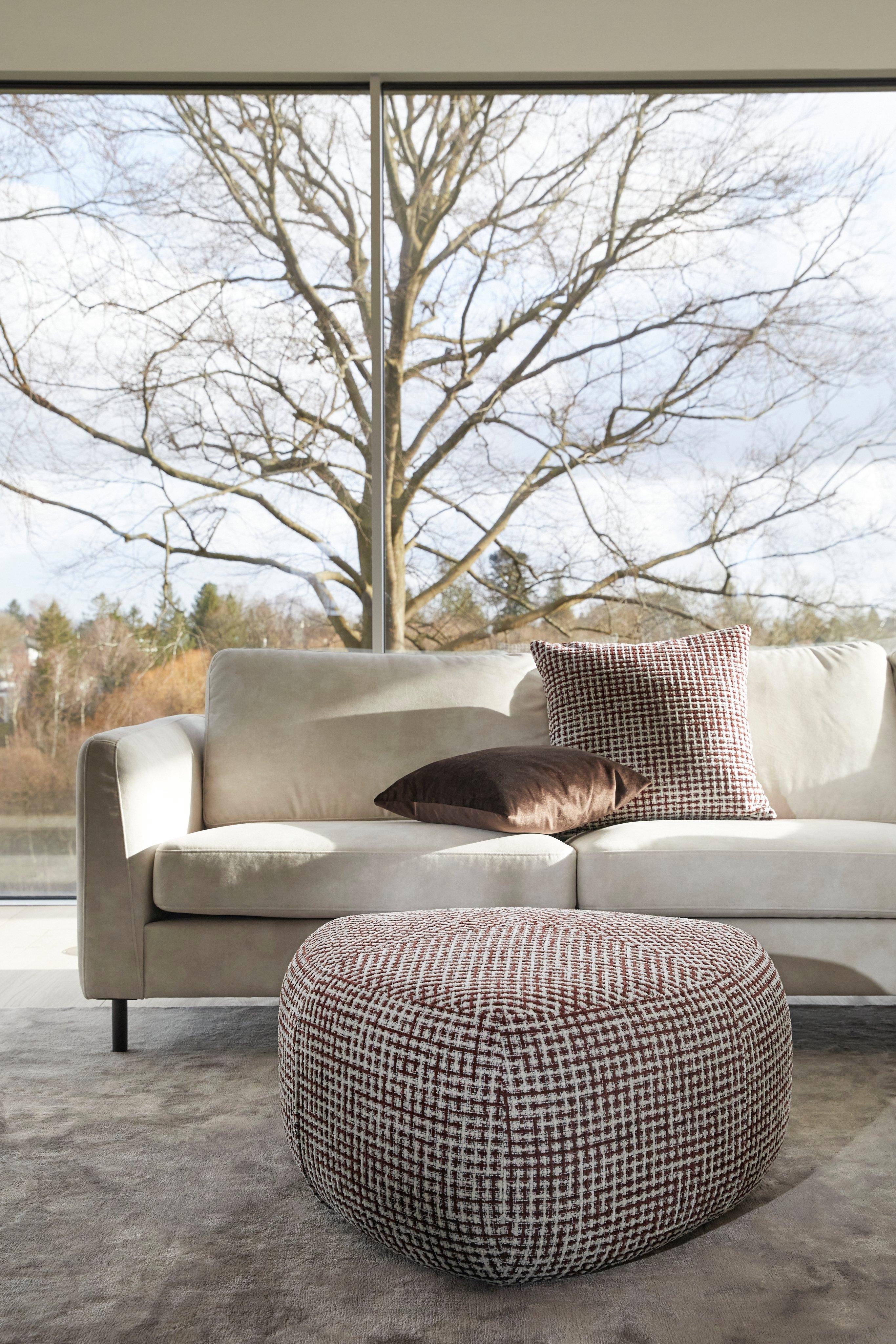 Sala moderna con sofá beige claro, cojines marrón y con estampado de cuadros y puf a cuadros a juego frente a una gran ventana con vista al árbol.