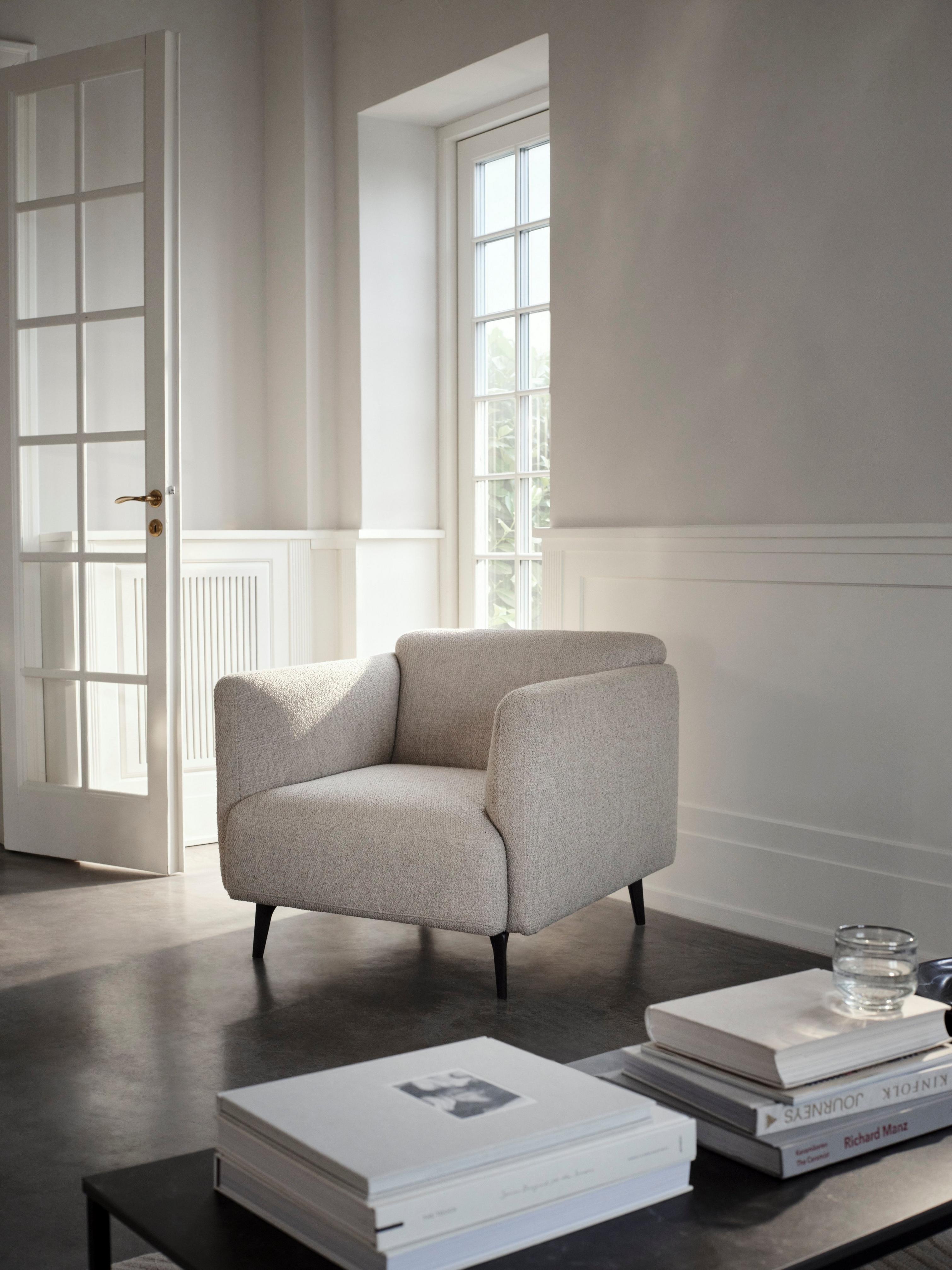 Fauteuil beige dans un salon moderne, près d'une fenêtre. Livres et verre sur la table basse.