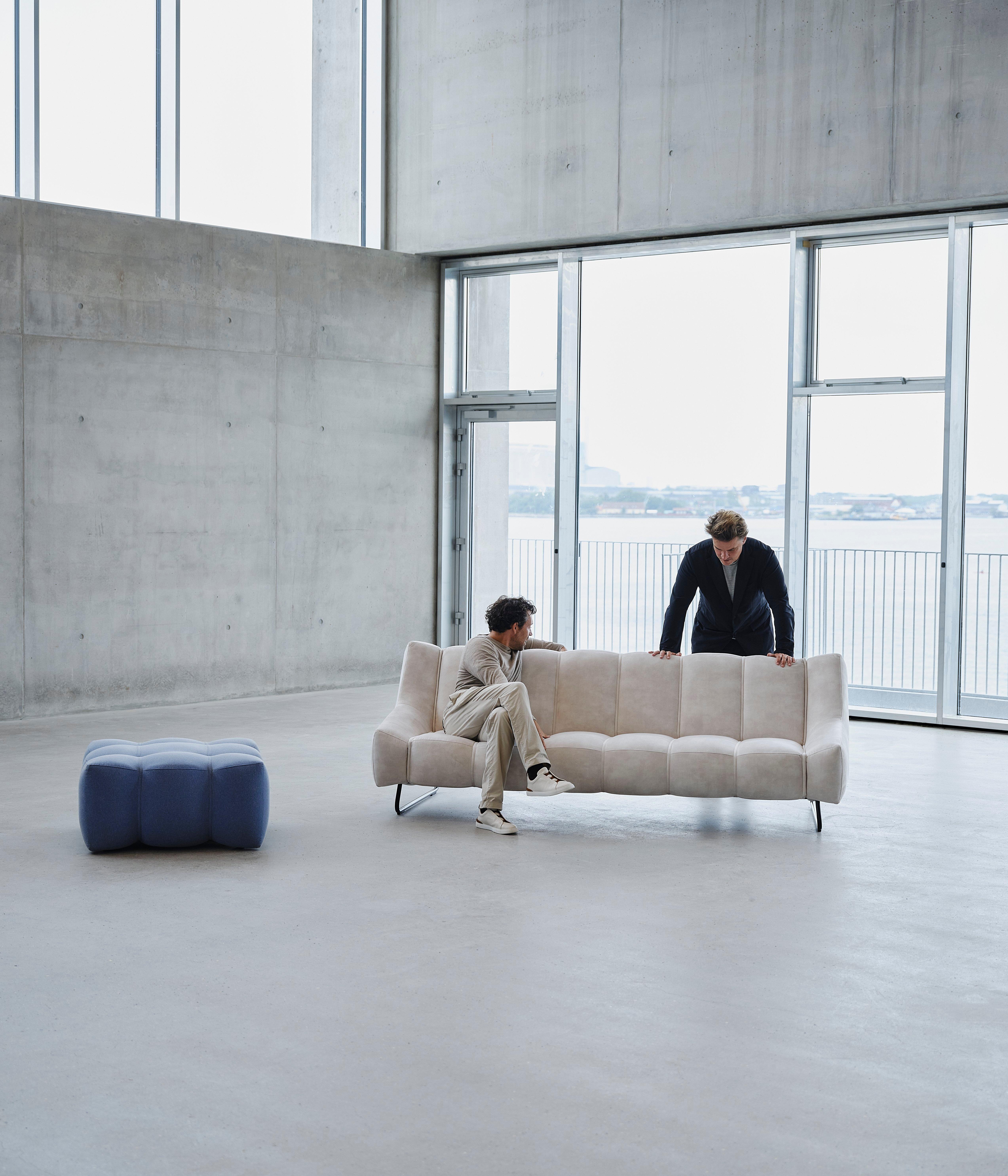 Two men interact with a light beige sofa in a spacious, minimalist room with concrete walls and large windows overlooking water.