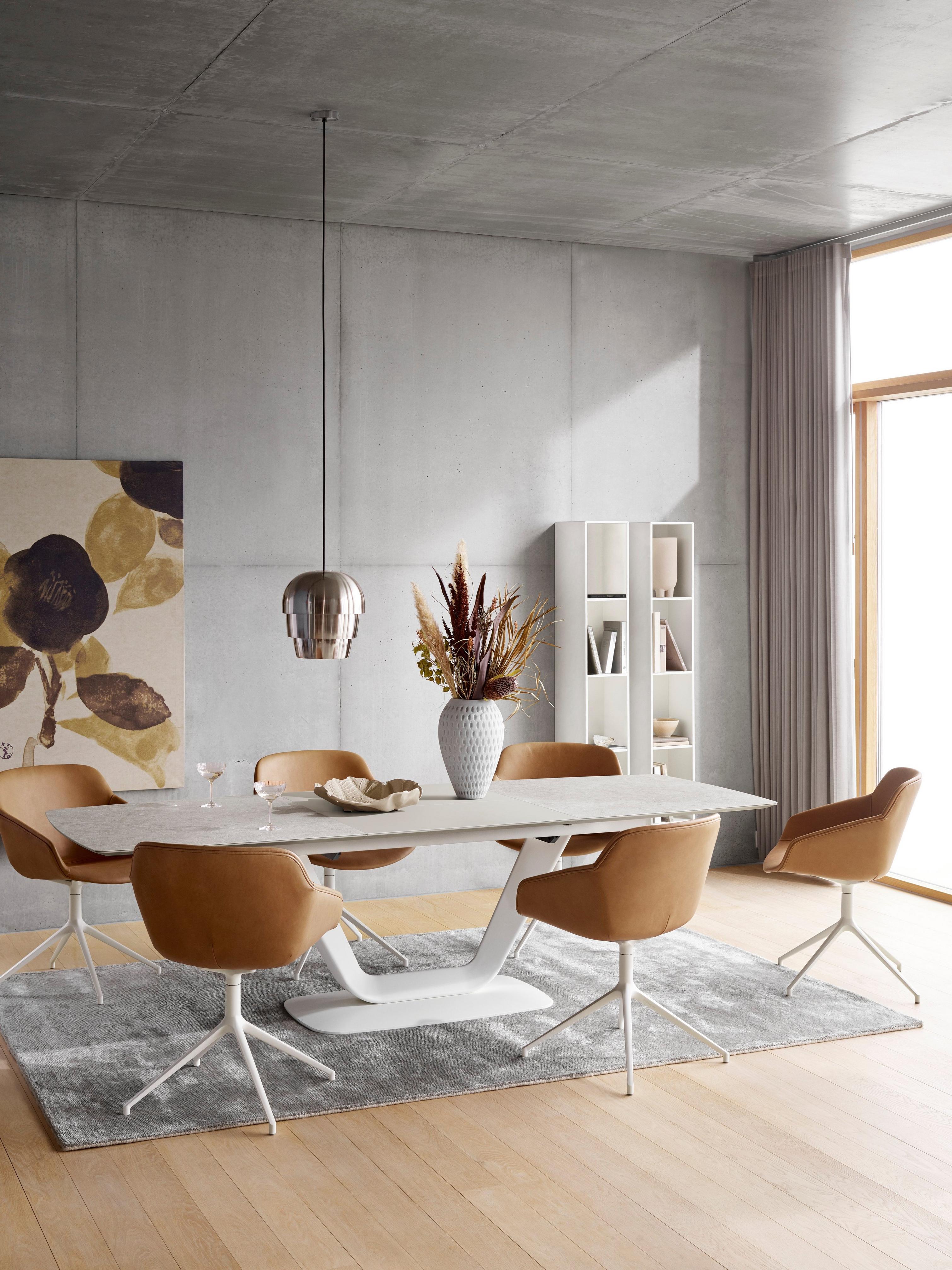Dining room with tan chairs, white table, pendant light, and gray rug, with art and shelving.