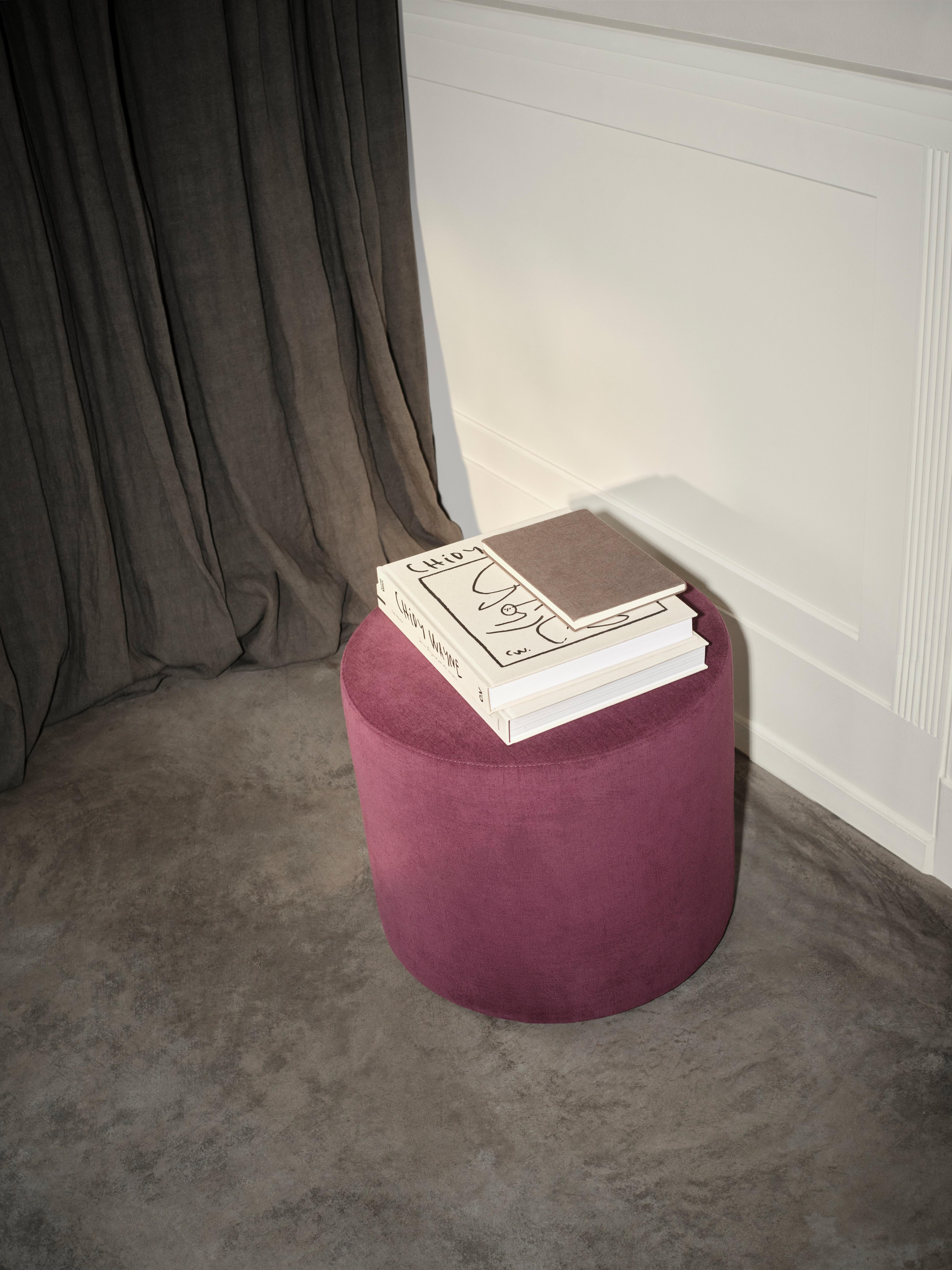 Round purple ottoman with stacked books on top, placed on a grey concrete floor beside dark curtains and white paneled wall.