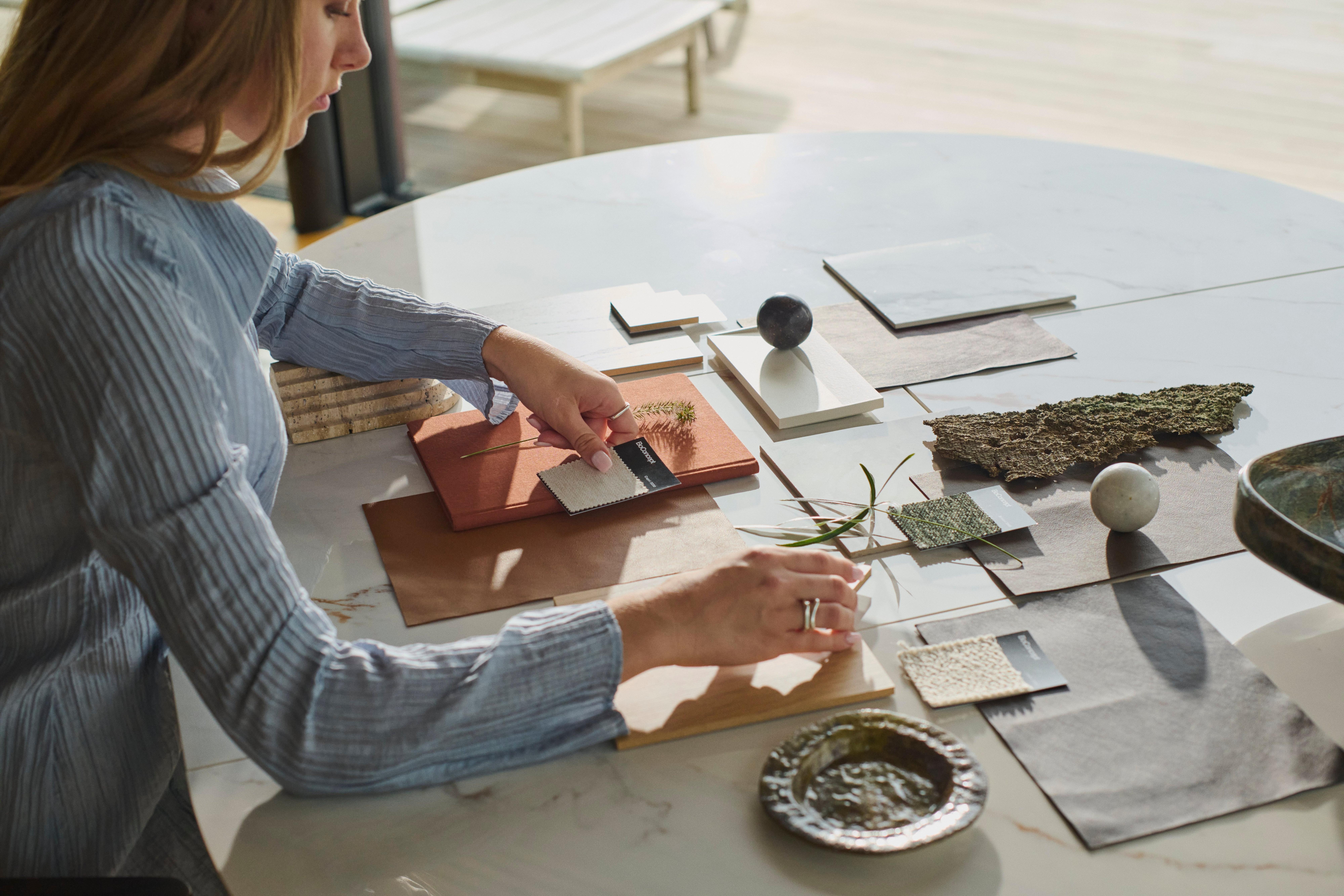 Una mujer selecciona muestras de telas y materiales en una mesa de mármol, organizando madera, textiles y acabados en piedra en una paleta de tonos neutros.