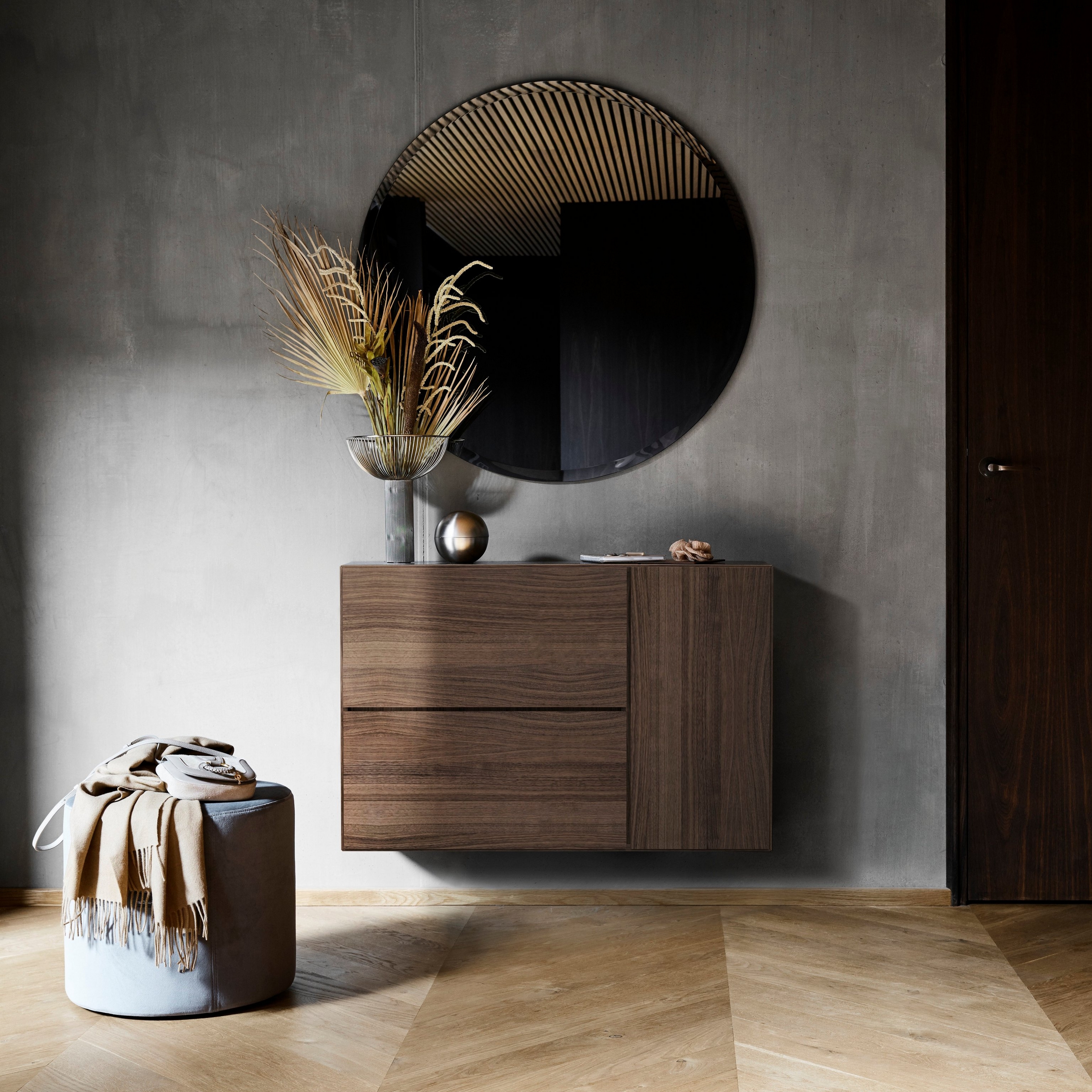 Stylish entryway with round mirror, wooden cabinet, vase with dried plants, and leather pouf.