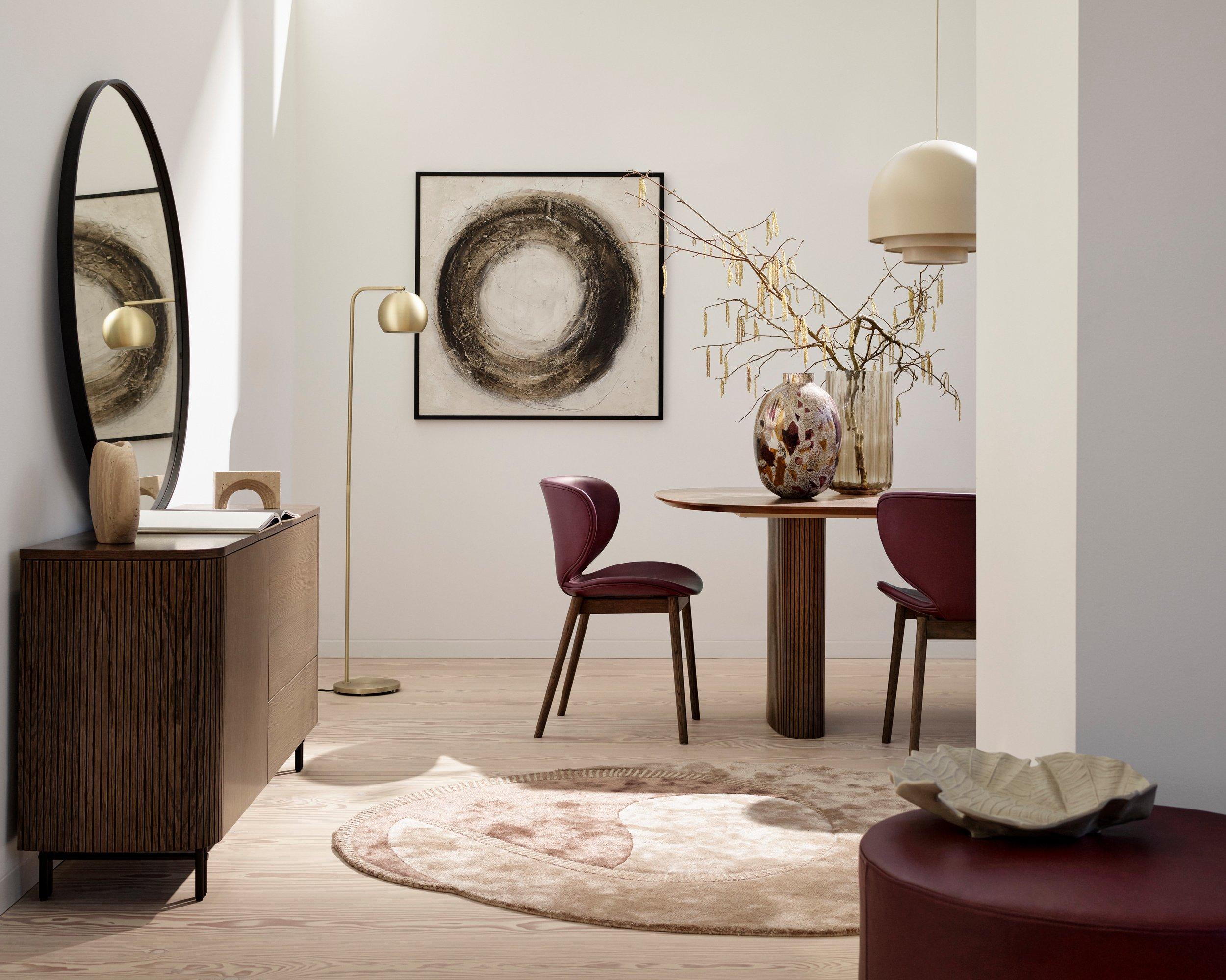 Minimalistická jedáleň s dvomi hnedými jedálenskými stoličkami Seoul, okrúhlym jedálenským stolom Santiago, abstraktným obrazom, príborníkom Santiago, stojanovou lampou a veľkými oknami.