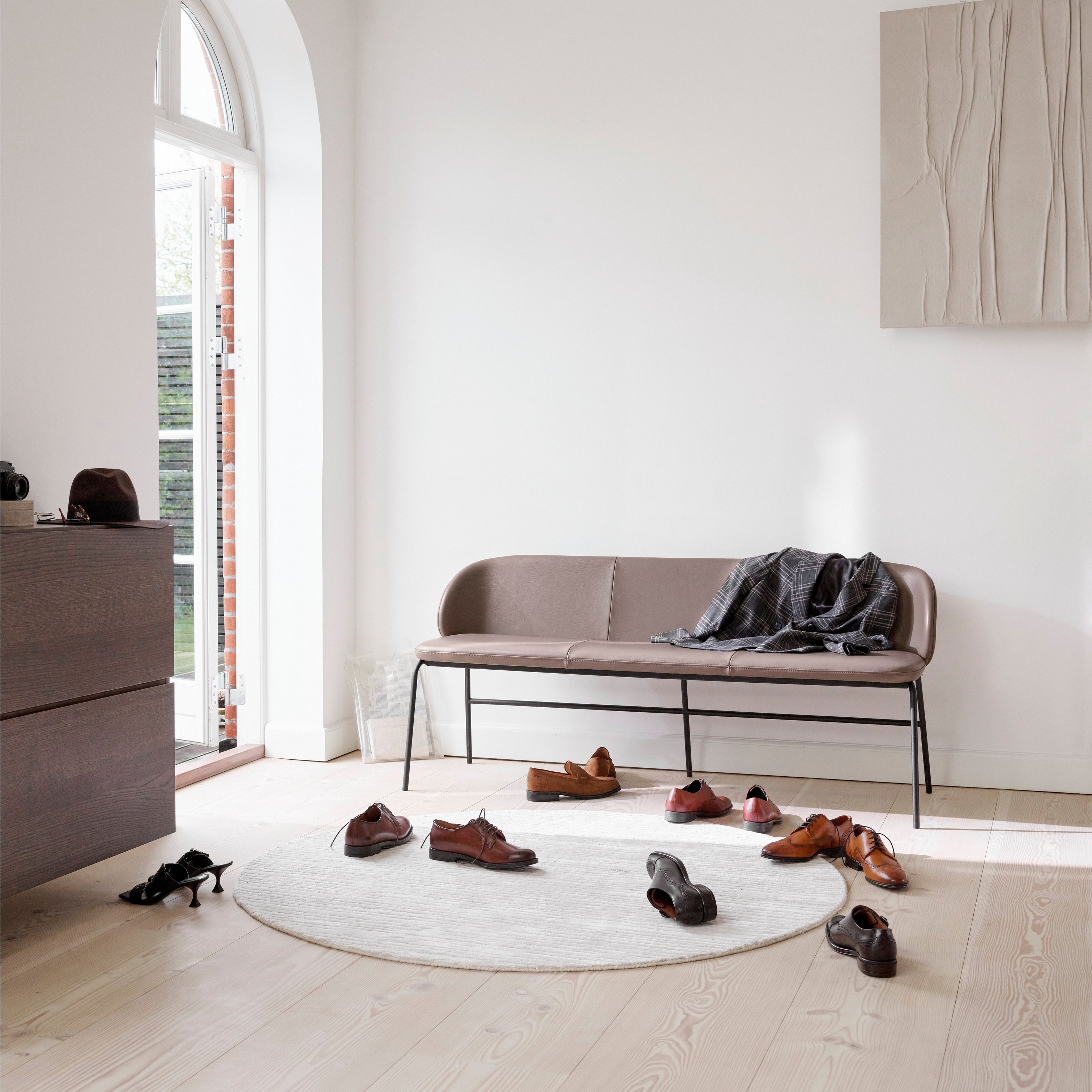 Kožená lavica v svetlej miestnosti s oblúkovitým oknom, topánkami na podlahe a dreveným umeleckým dielom.