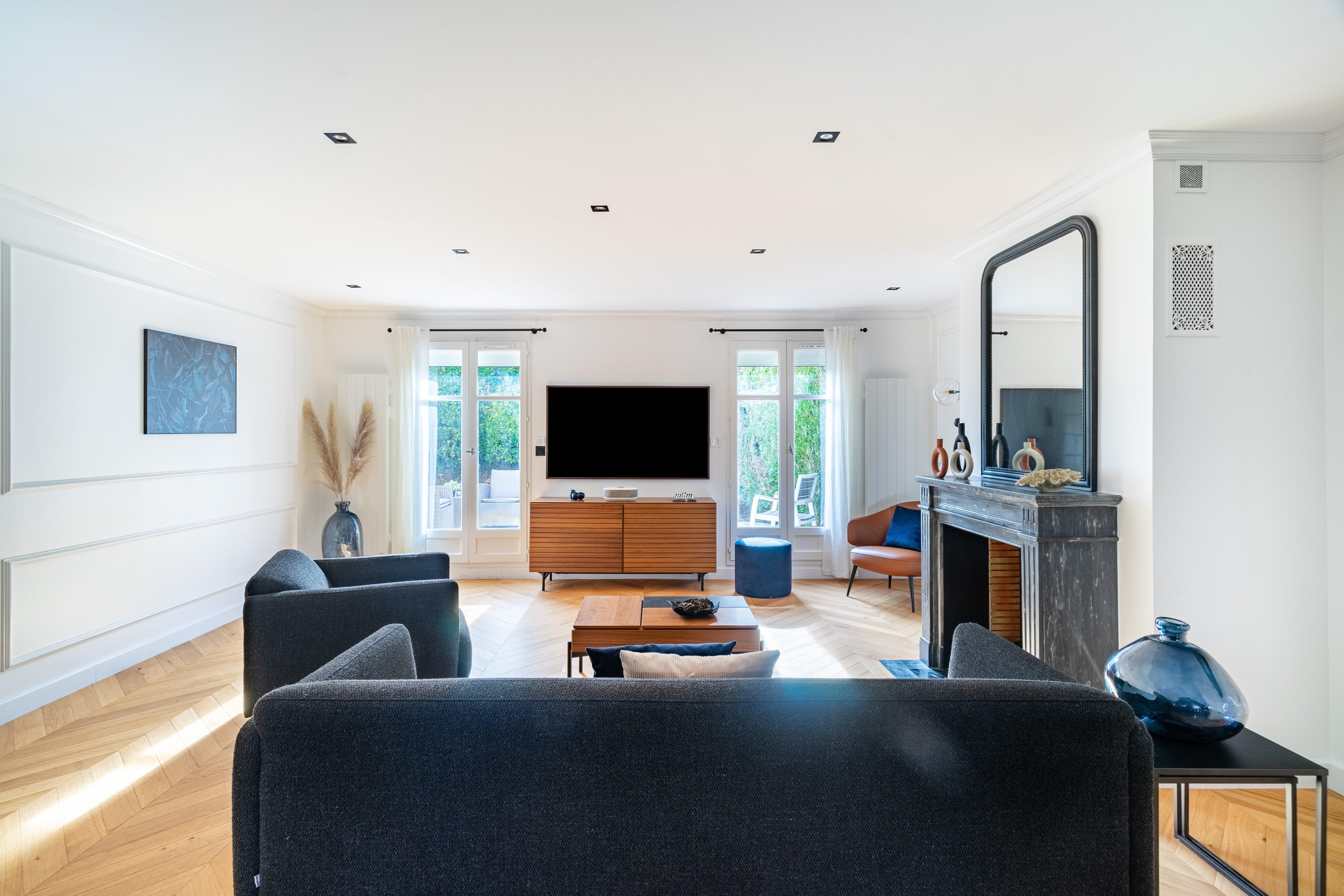 Symmetrical living room view with dark sofas, fireplace, TV console, and large windows framed by white curtains and greenery.