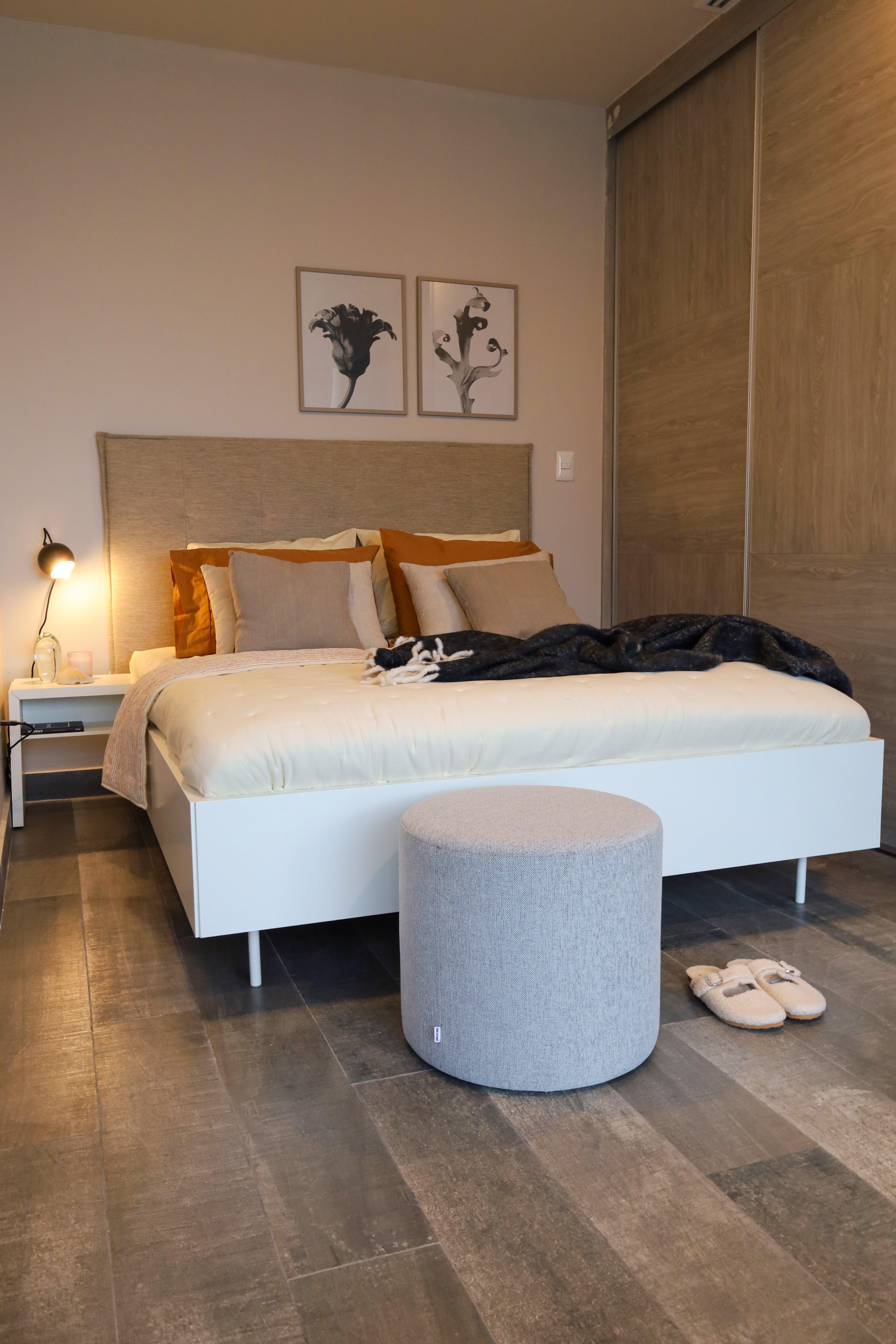 Cosy bedroom with a white bed, grey ottoman, slippers, and wall art above the headboard.