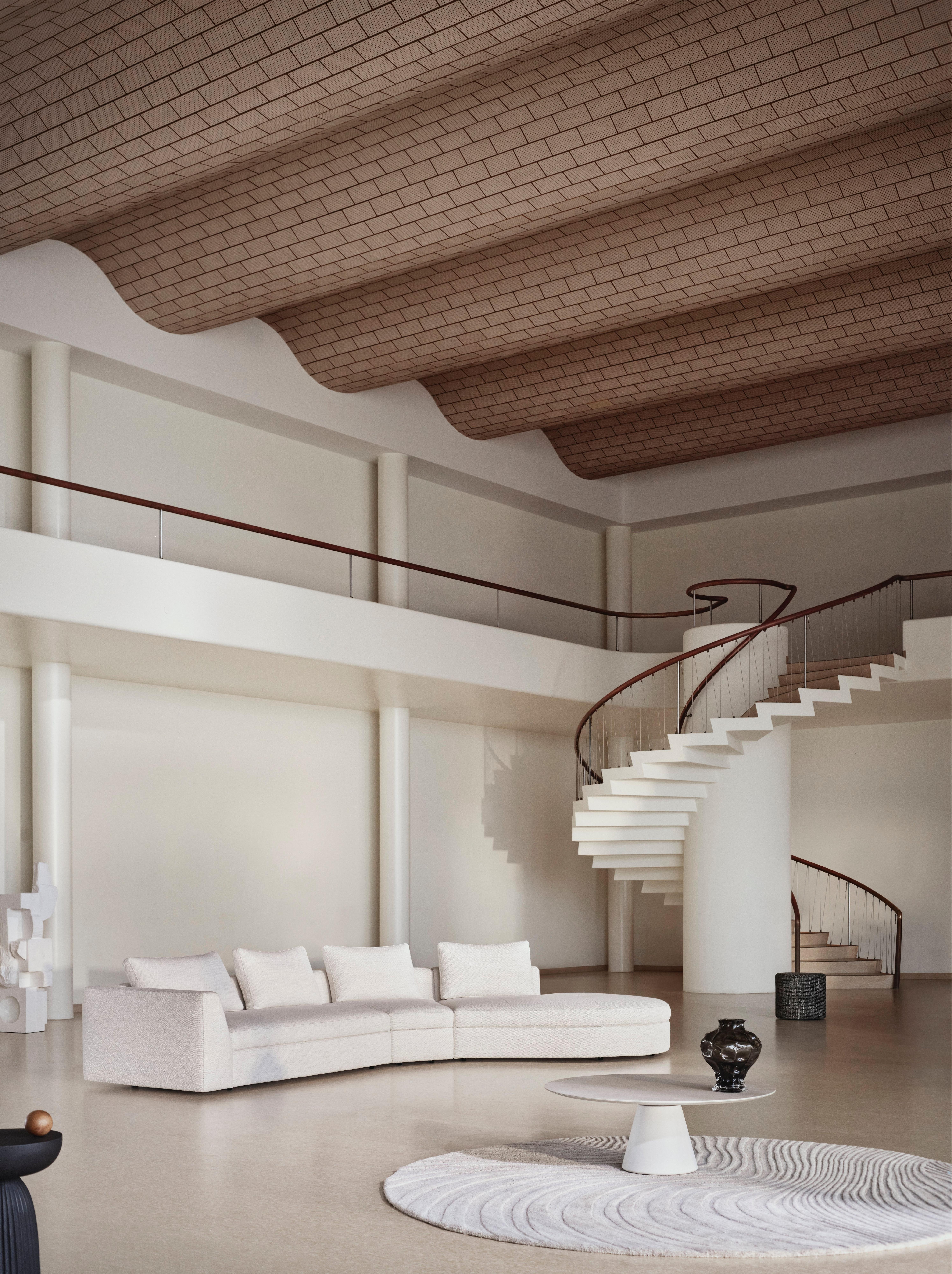 Spacious interior with a curved white sectional sofa, sculptural spiral staircase, round rug, and high ceiling with arched brick pattern.