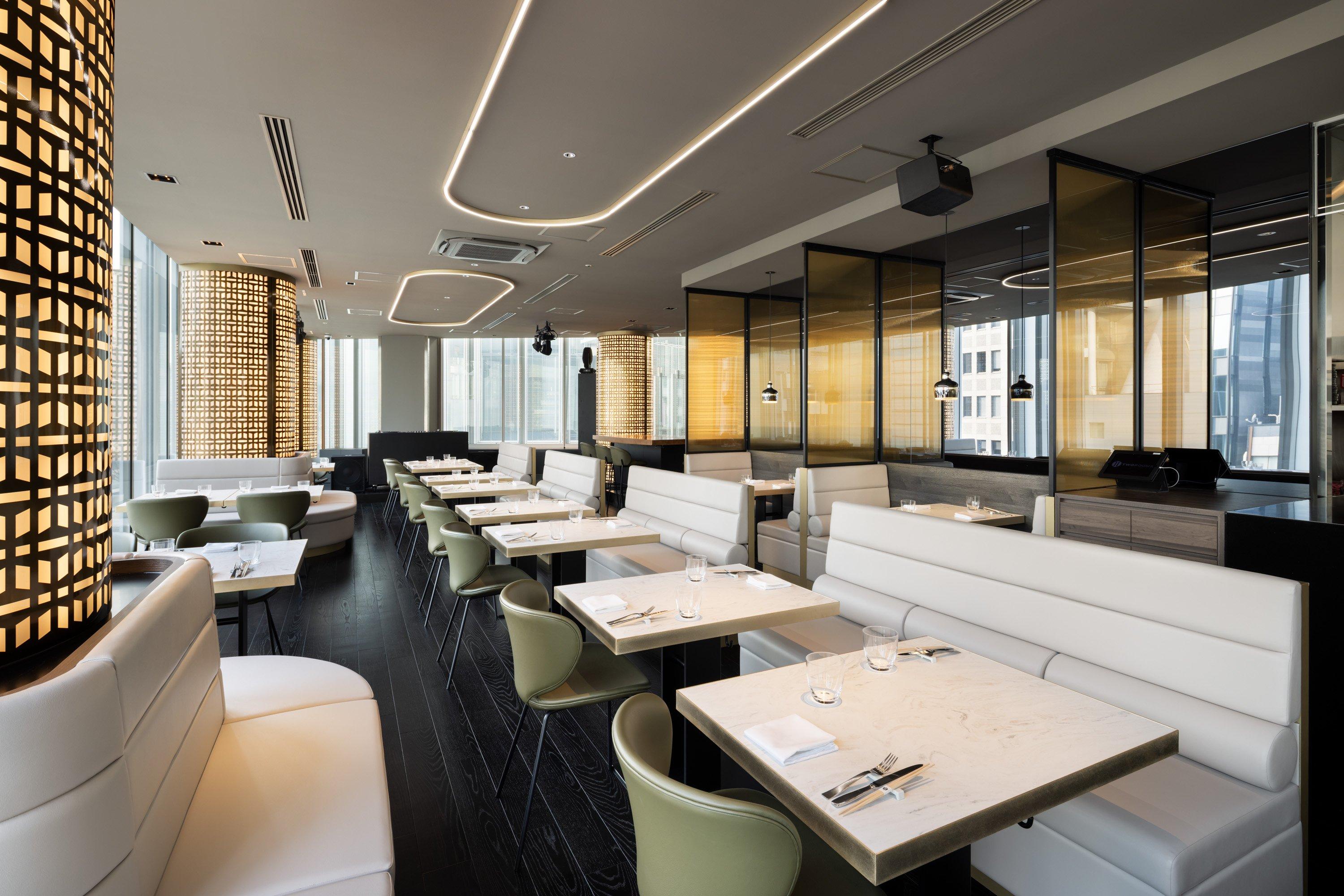 Restaurante moderno con mesas y sillas blancas, luz cálida y ventanales grandes.