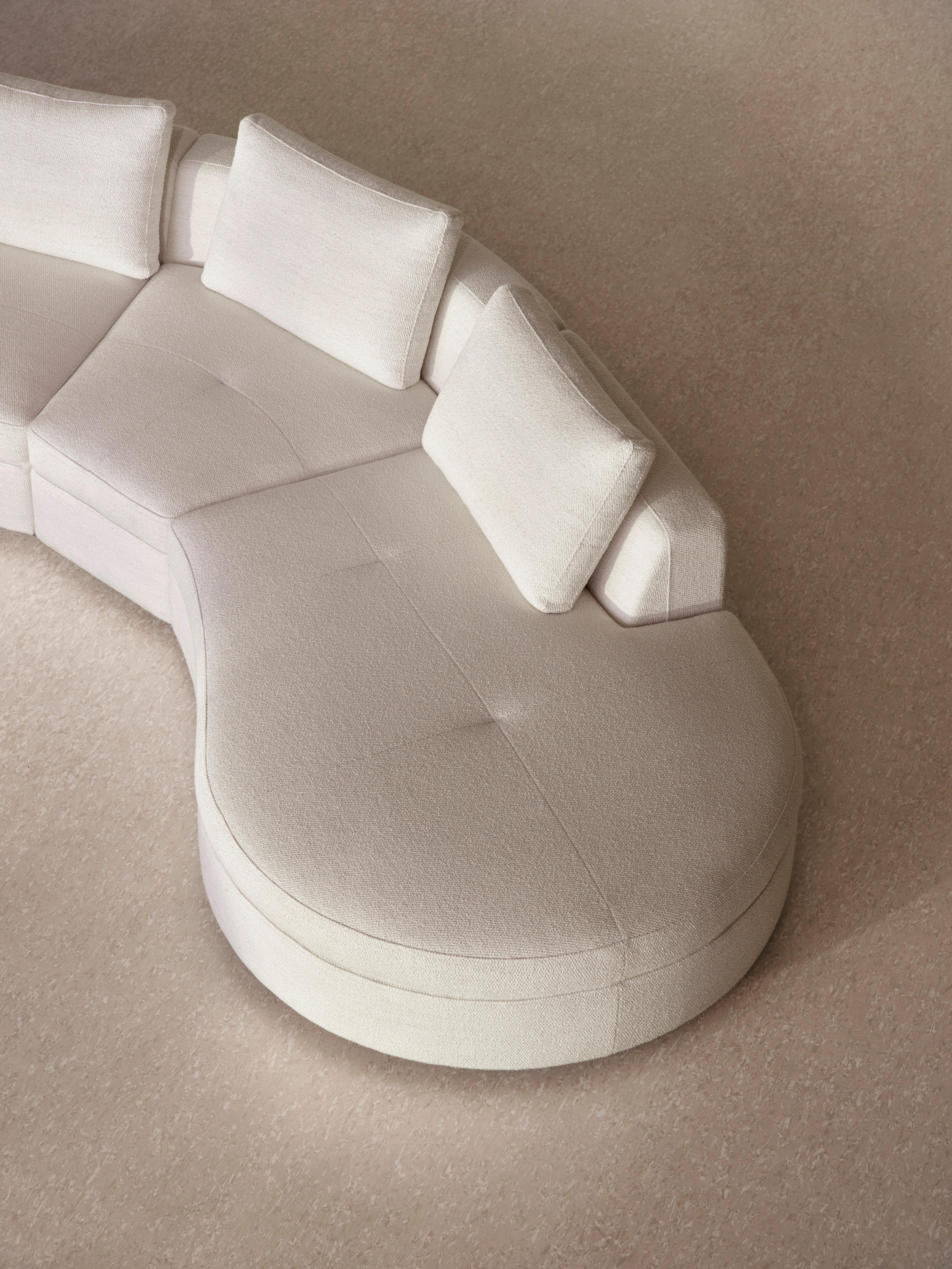 Sofá modular curvo Bergamo em tecido claro com três almofadas para as costas, colocado num pavimento com textura em tons neutros.