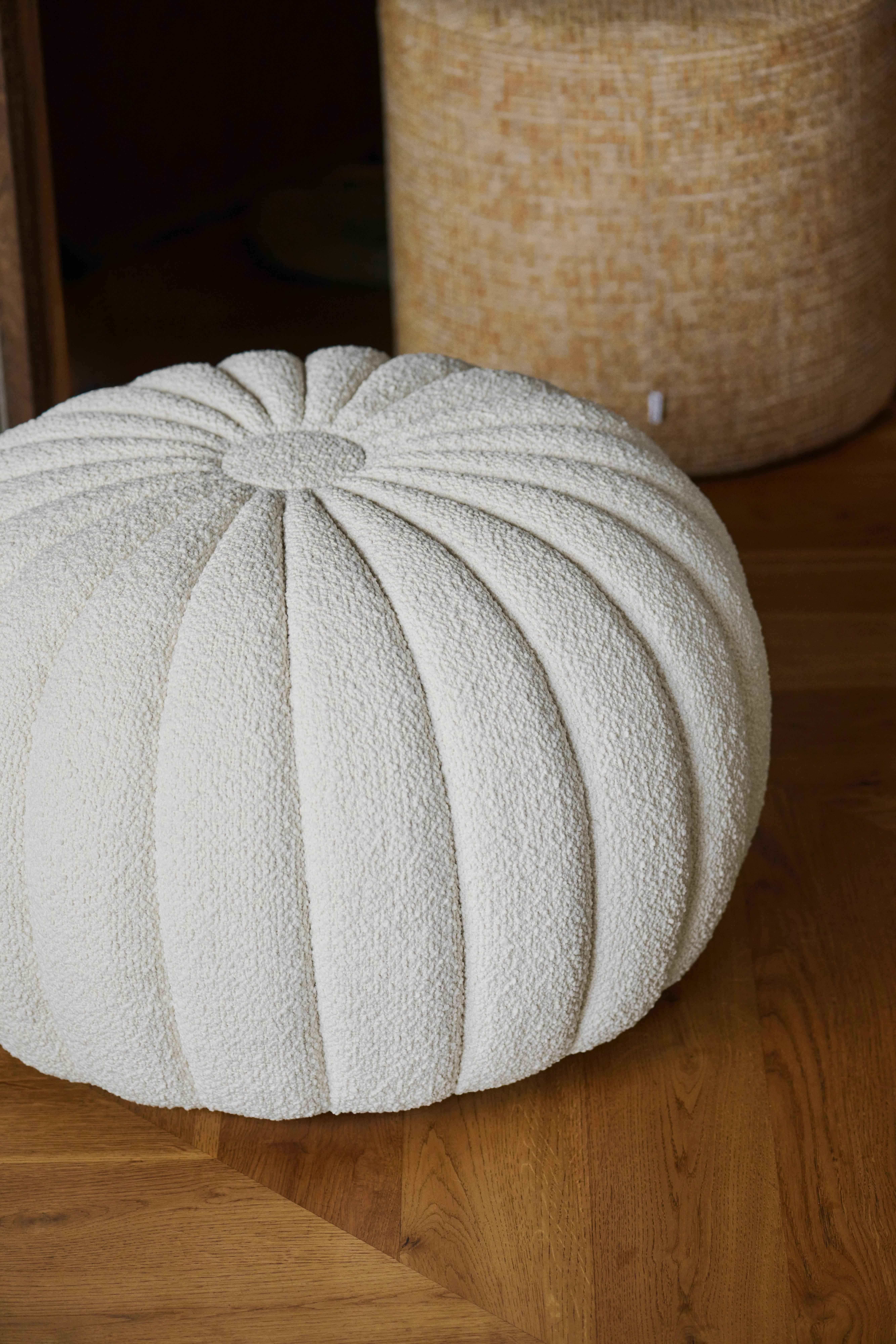 Vit rund puff med ribbad textur på trägolv. Brun cylinderformad puff i bakgrunden.