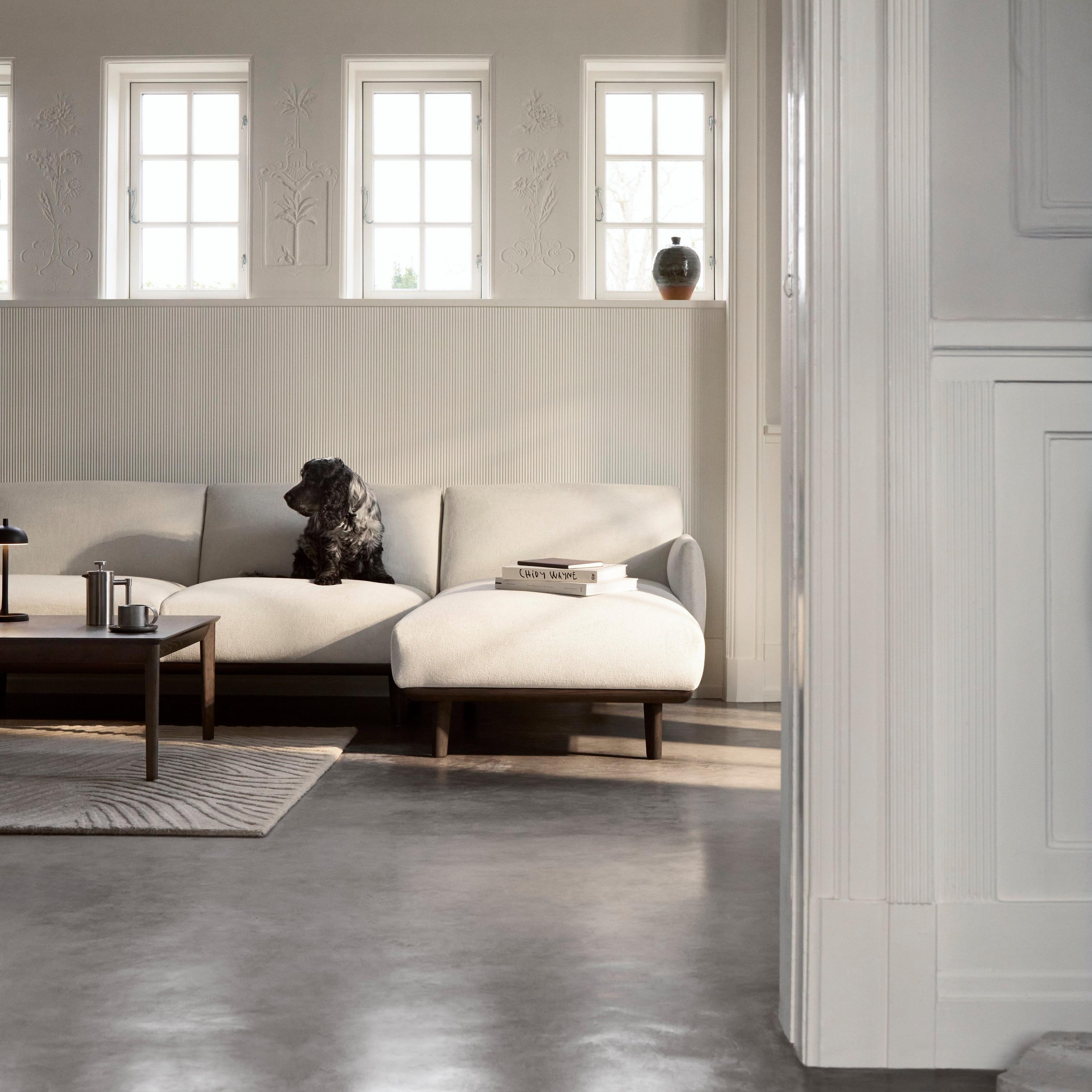 Aarhus sofa in light upholstery with slim wooden legs, set in a classic room with white paneling, grid windows, and dark coffee table.