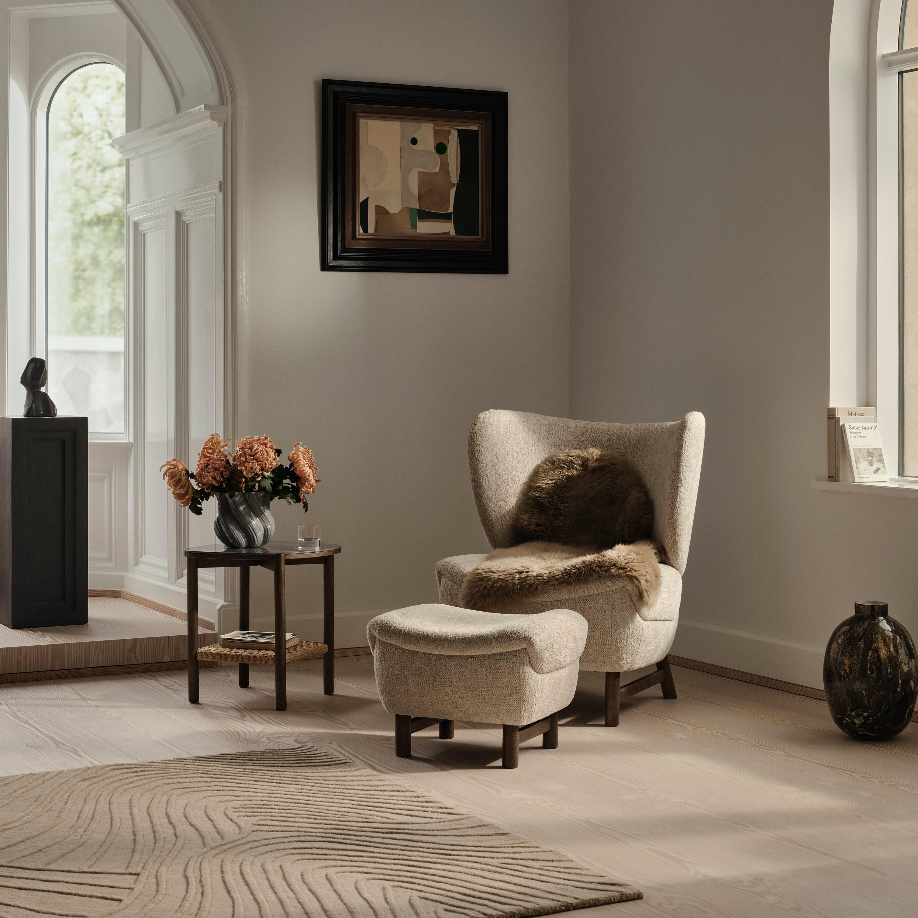 Cosy room with an armchair, footstool, side table with flowers, and large window.