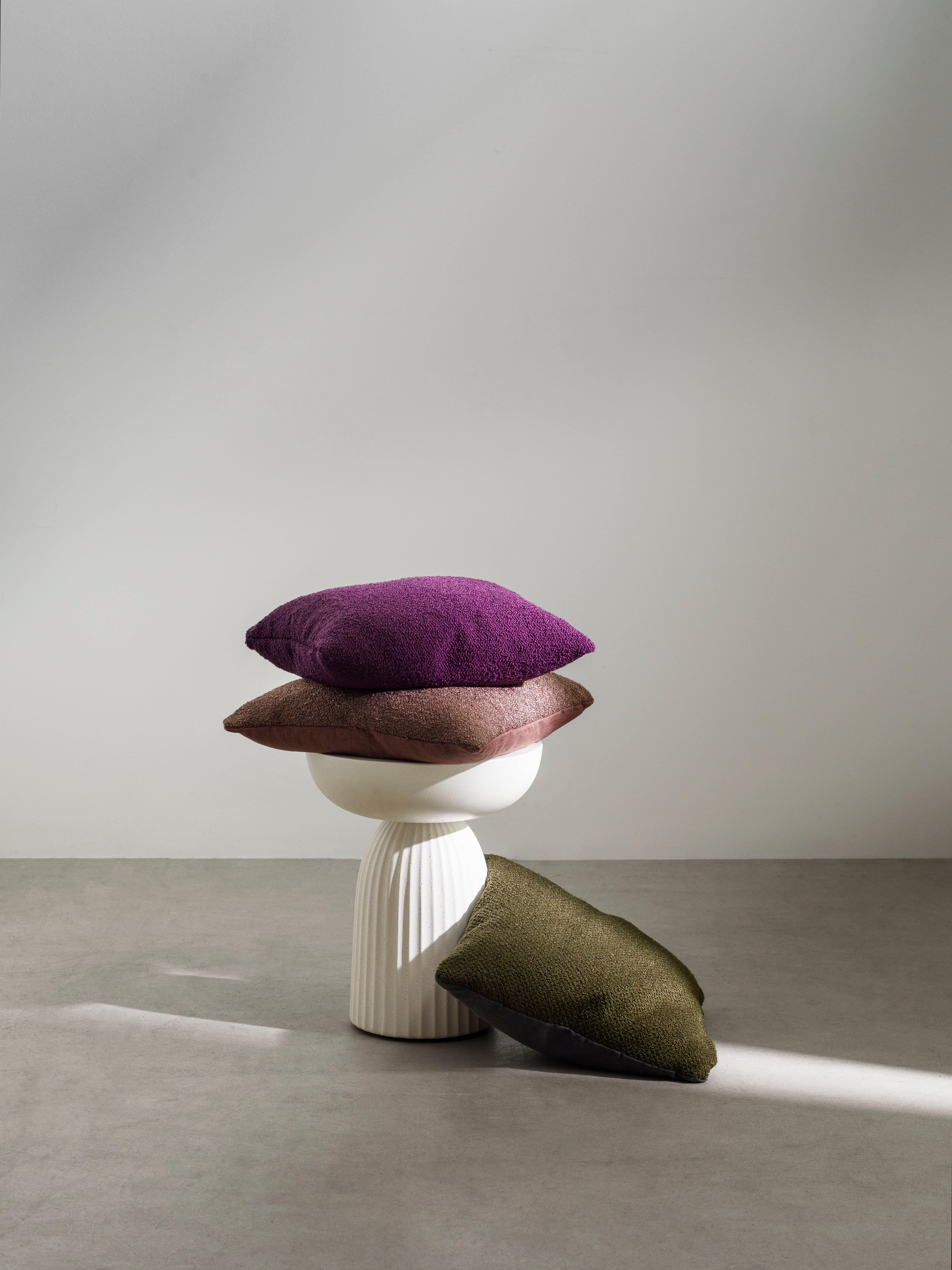 Tre teksturerte puter i lilla, brun og olivengrønn stablet på og ved siden av en hvit riflet sokkel mot en nøytral bakgrunn.
