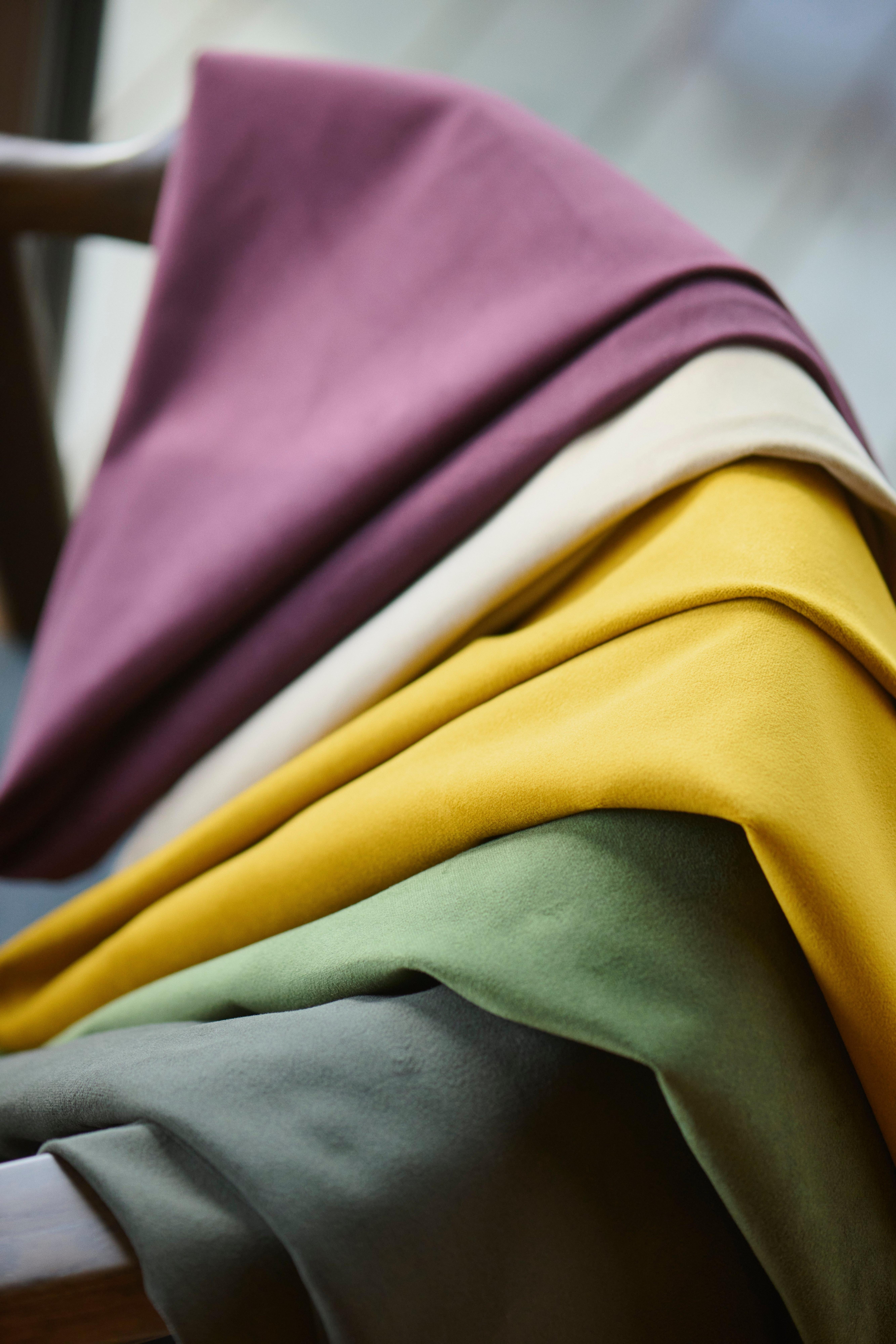 Puha szövetek visszafogott lila, krémszínű, mustársárga, zöld és szürke árnyalatokban, fa székre halmozva.