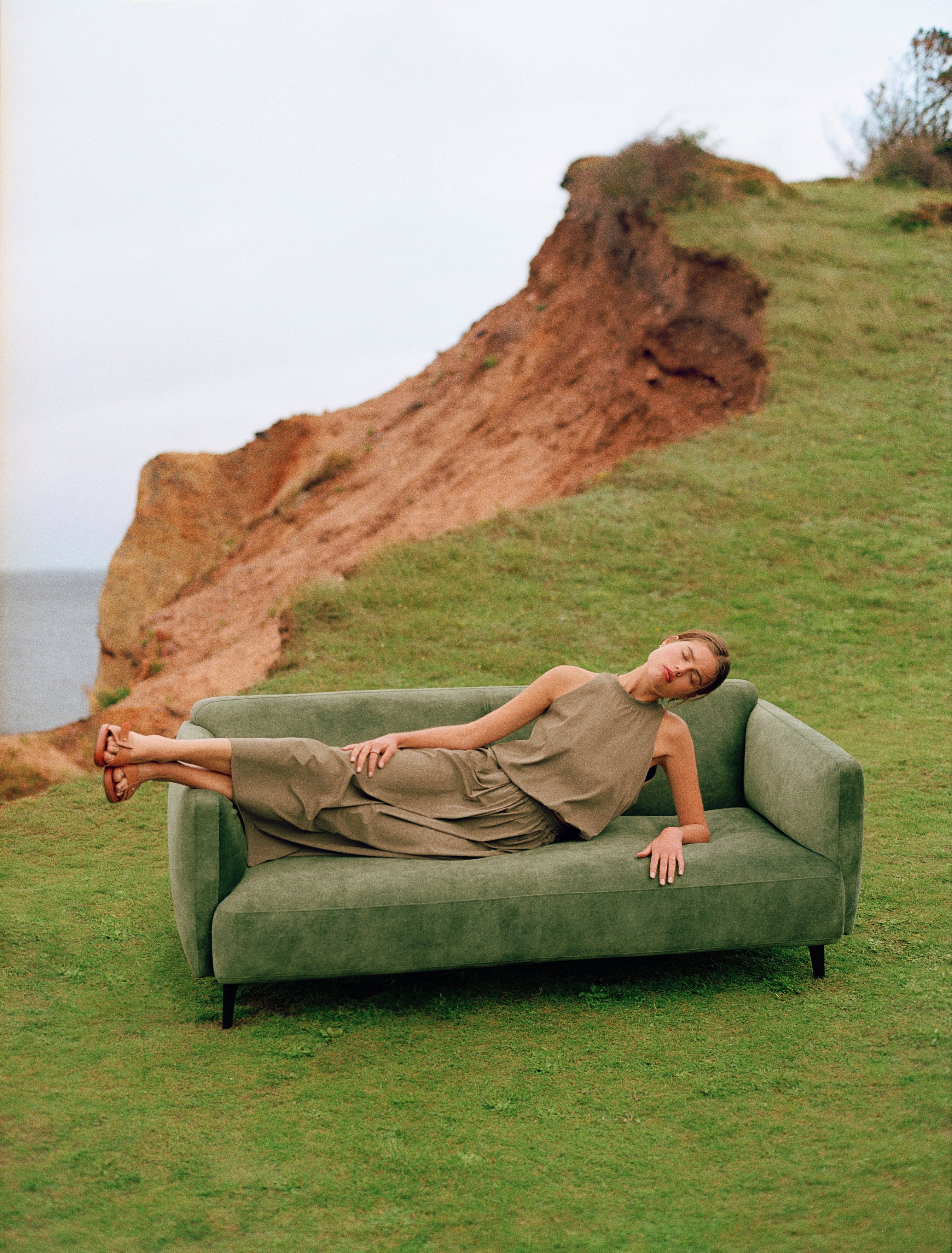 Woman reclining on a Modena sofa in a grassy outdoor setting with coastal cliffs.