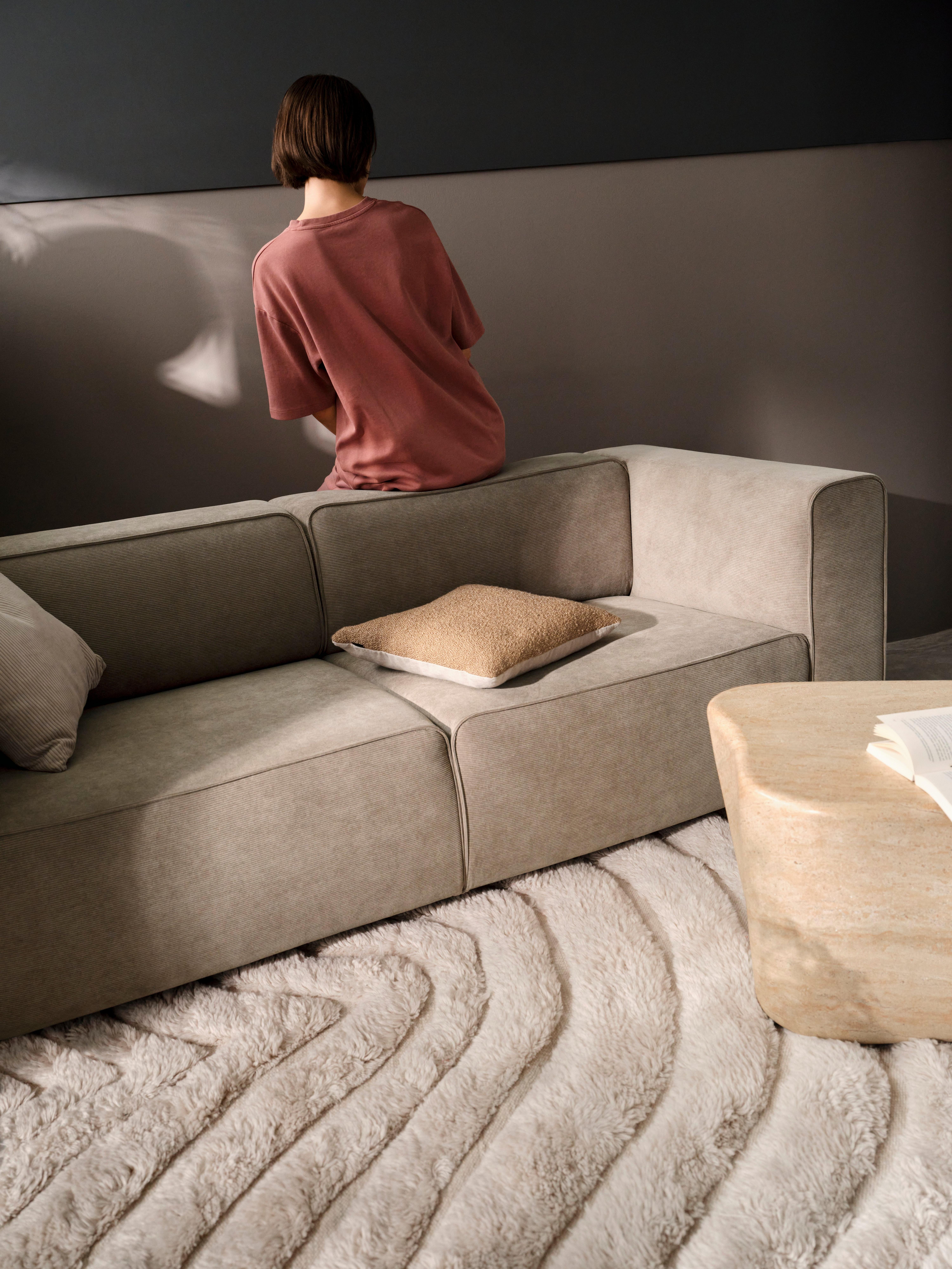 Person in a pink oversized t-shirt sits on the back of a beige sofa with a textured rug, facing a two-tone wall. A beige cushion is on the sofa.