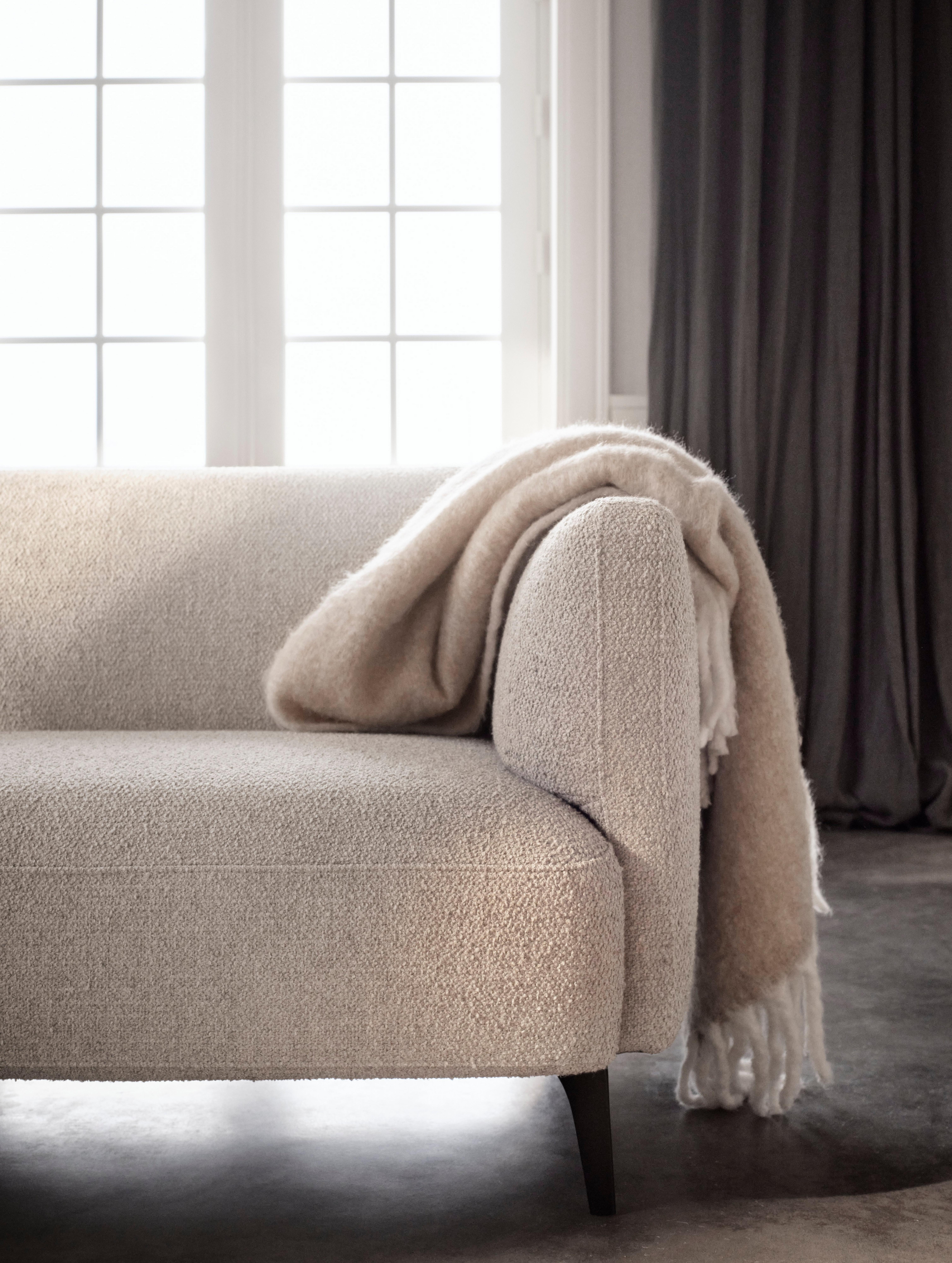 Modena sofa in beige textured fabric with curved armrest, dark wood legs, and a cream throw draped over the side.