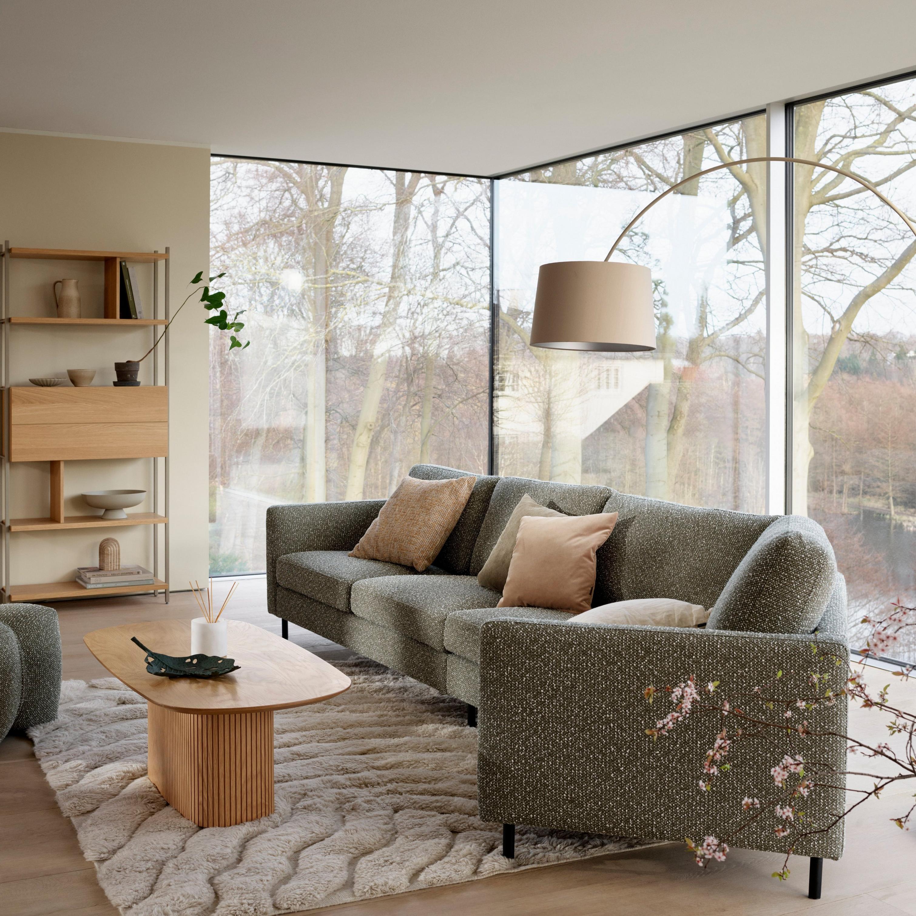 Modern living room with a green Indivi sofa and a light wood Santiago coffee table on a textured rug. Floor-to-ceiling windows offer a view of trees.