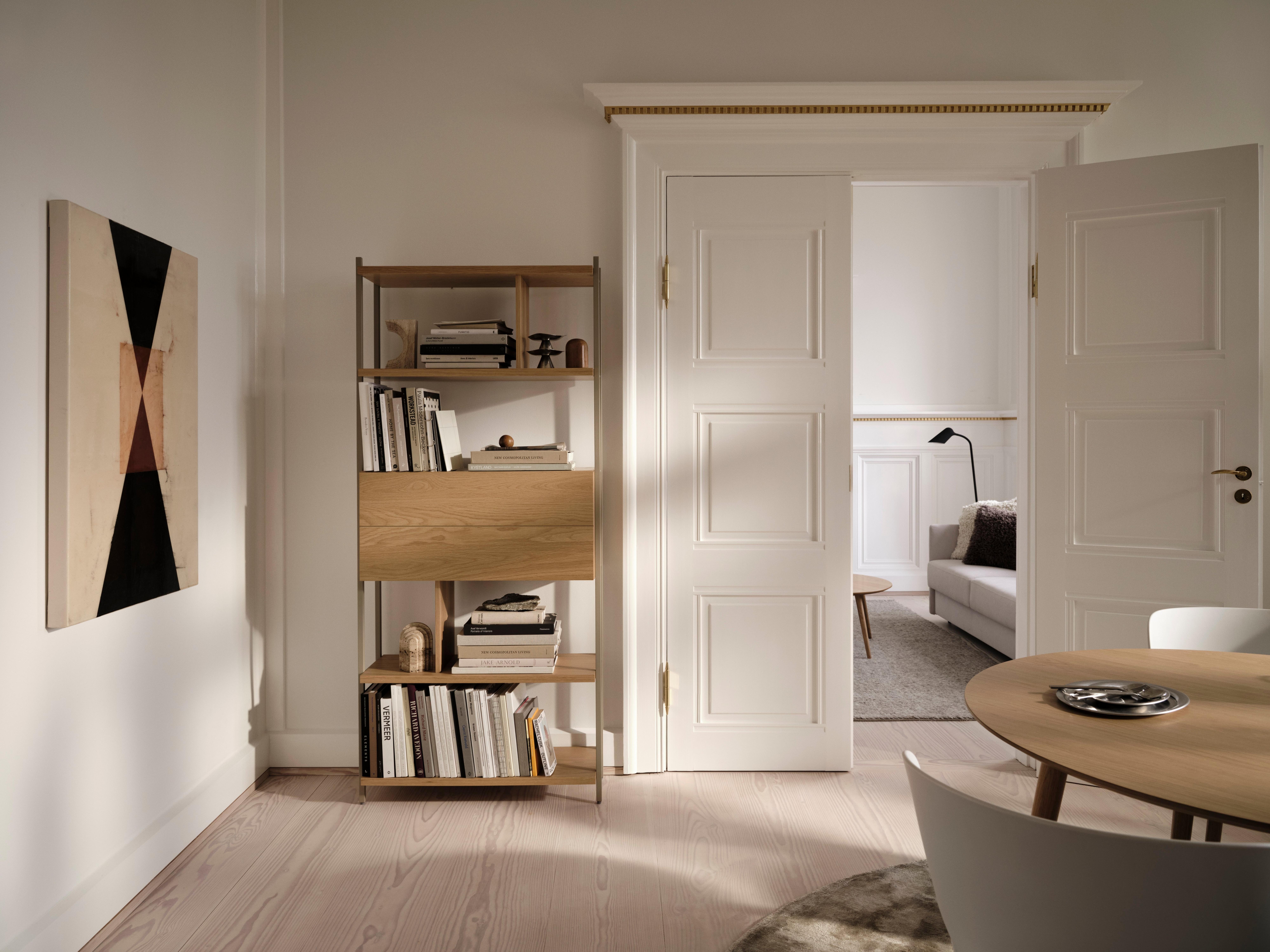 Lyst stue med træbord, hylde med bøger og kunst på væggen. Åben dør til sofaområde.