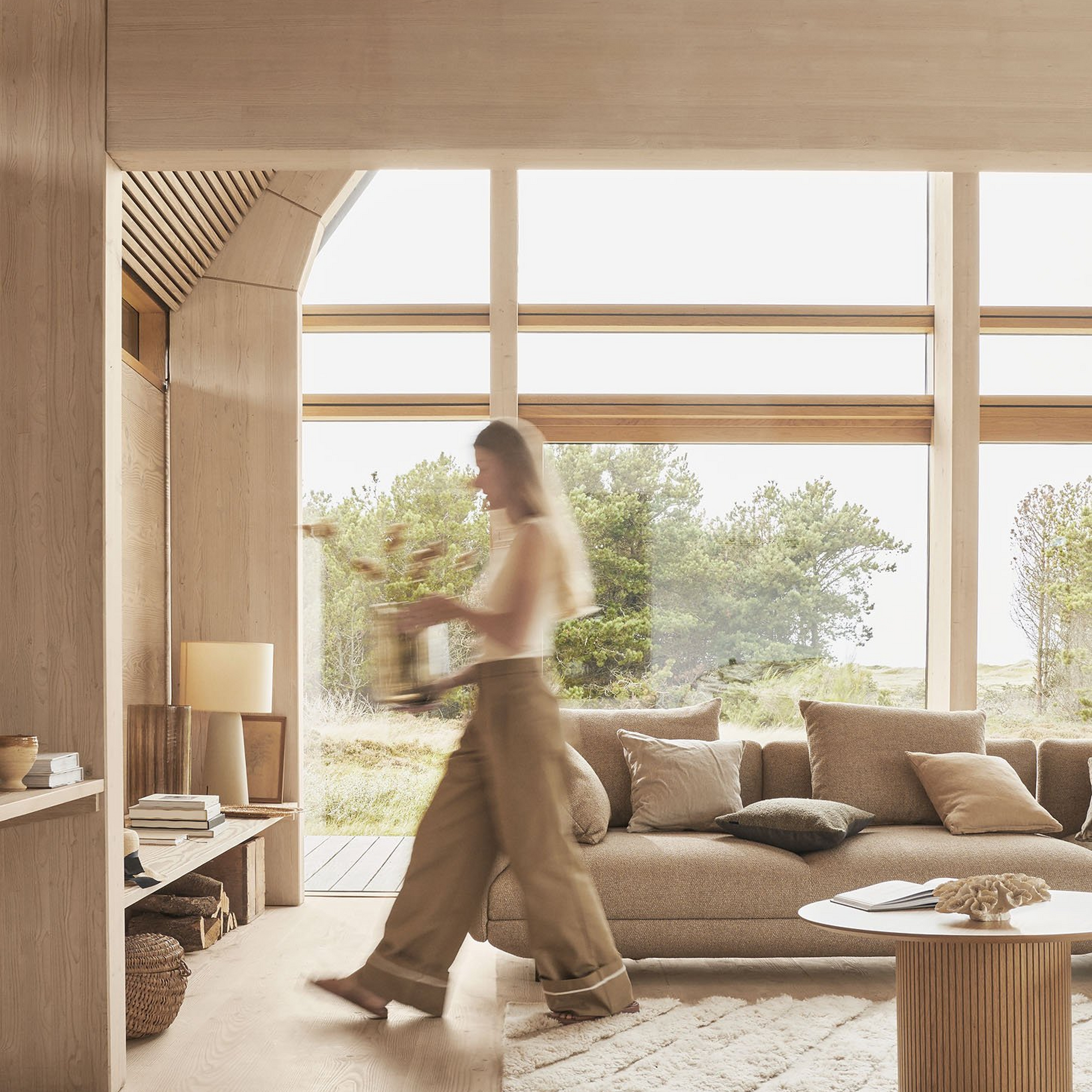 木材の梁を持つ日当たりの良い部屋に快適なソファが置かれ、動いている人物の姿がぼやけて写っている。