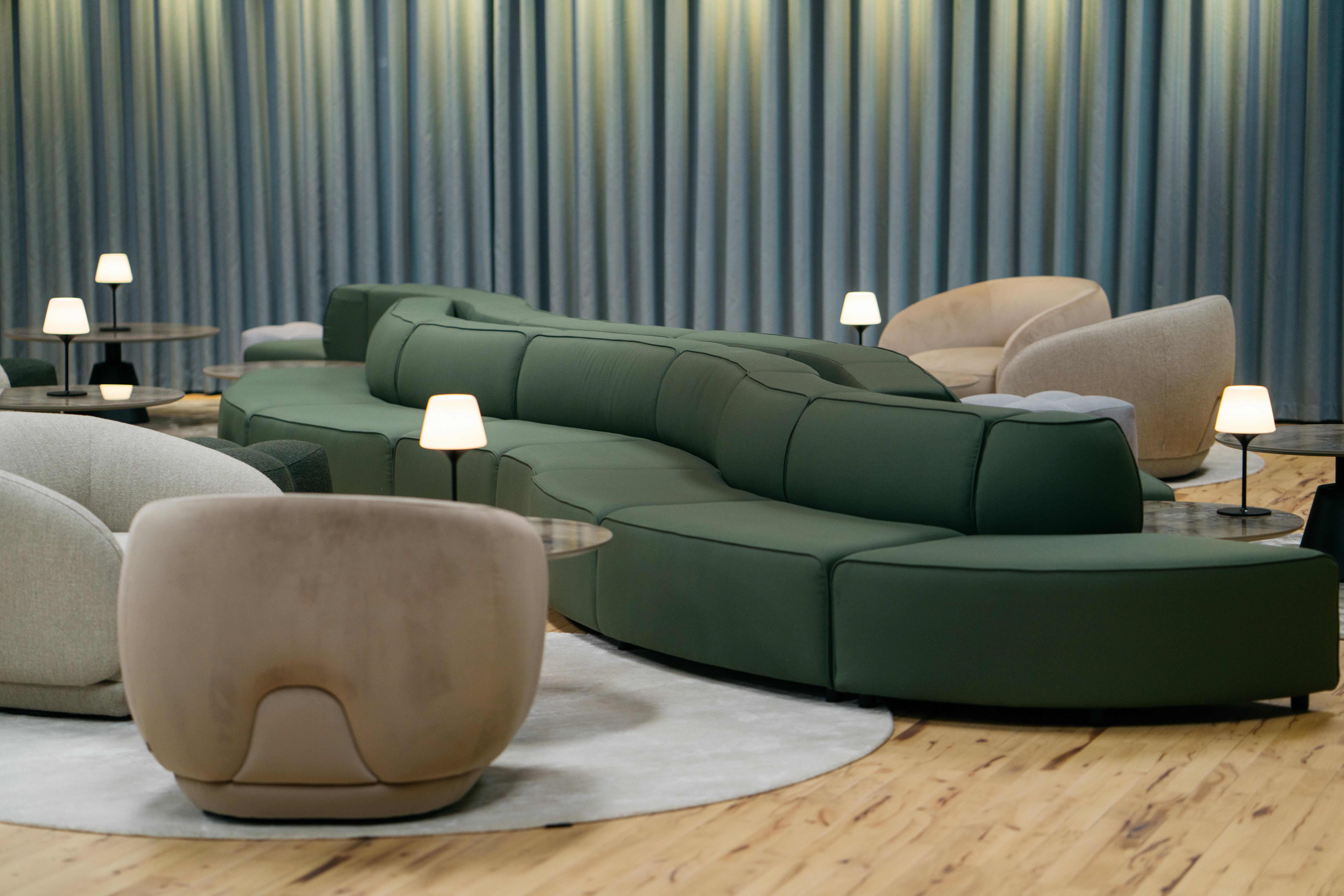 弧形深綠色 Carmo 蛇形沙發搭配米色圓形 Bolzano 扶手椅、小圓桌和柔軟桌燈。木頭地板和垂墜窗簾。