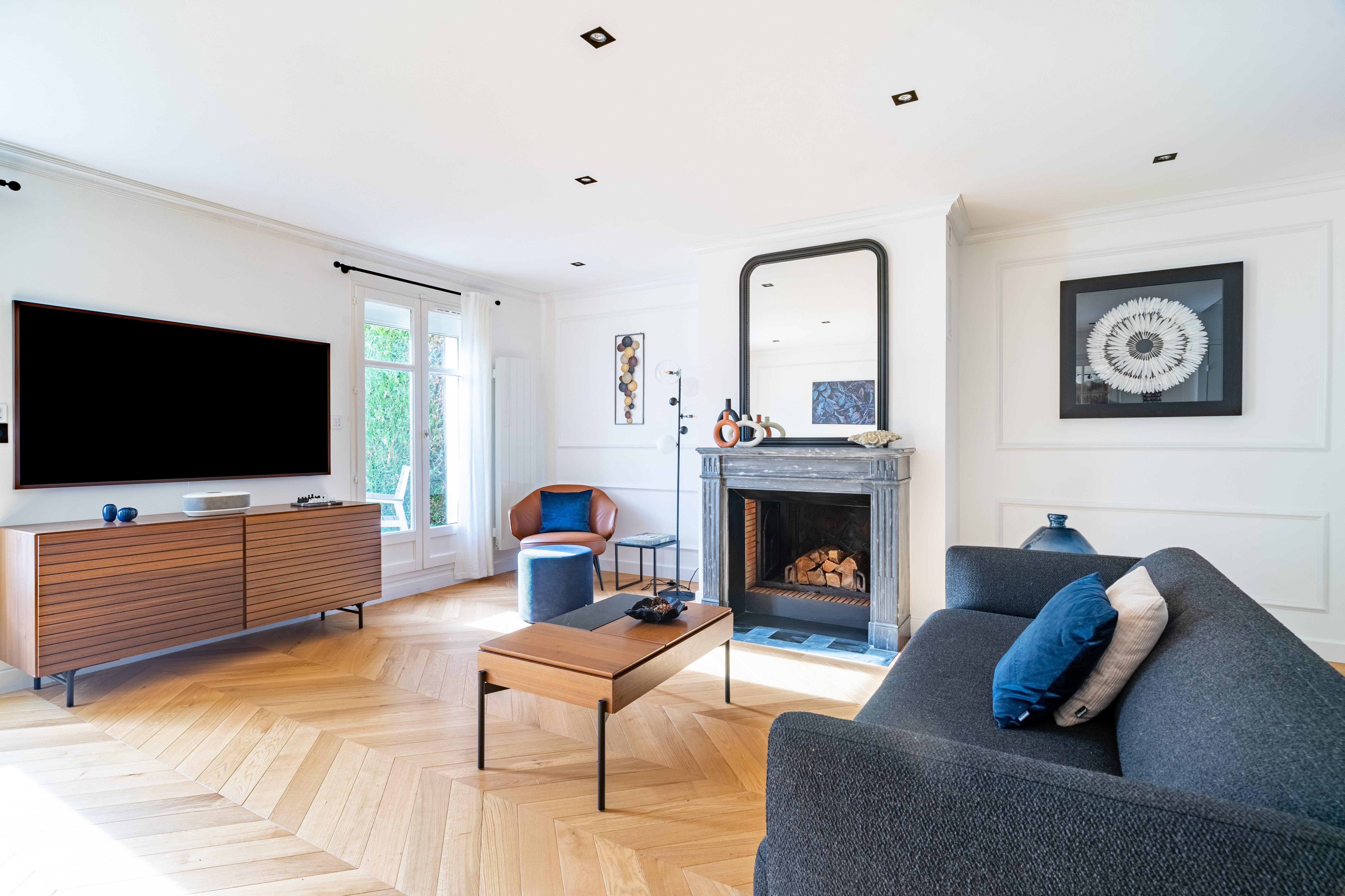 Bright living room with dark grey sofa, tan leather chair, wall-mounted TV, fireplace, and chevron-patterned wood floor.