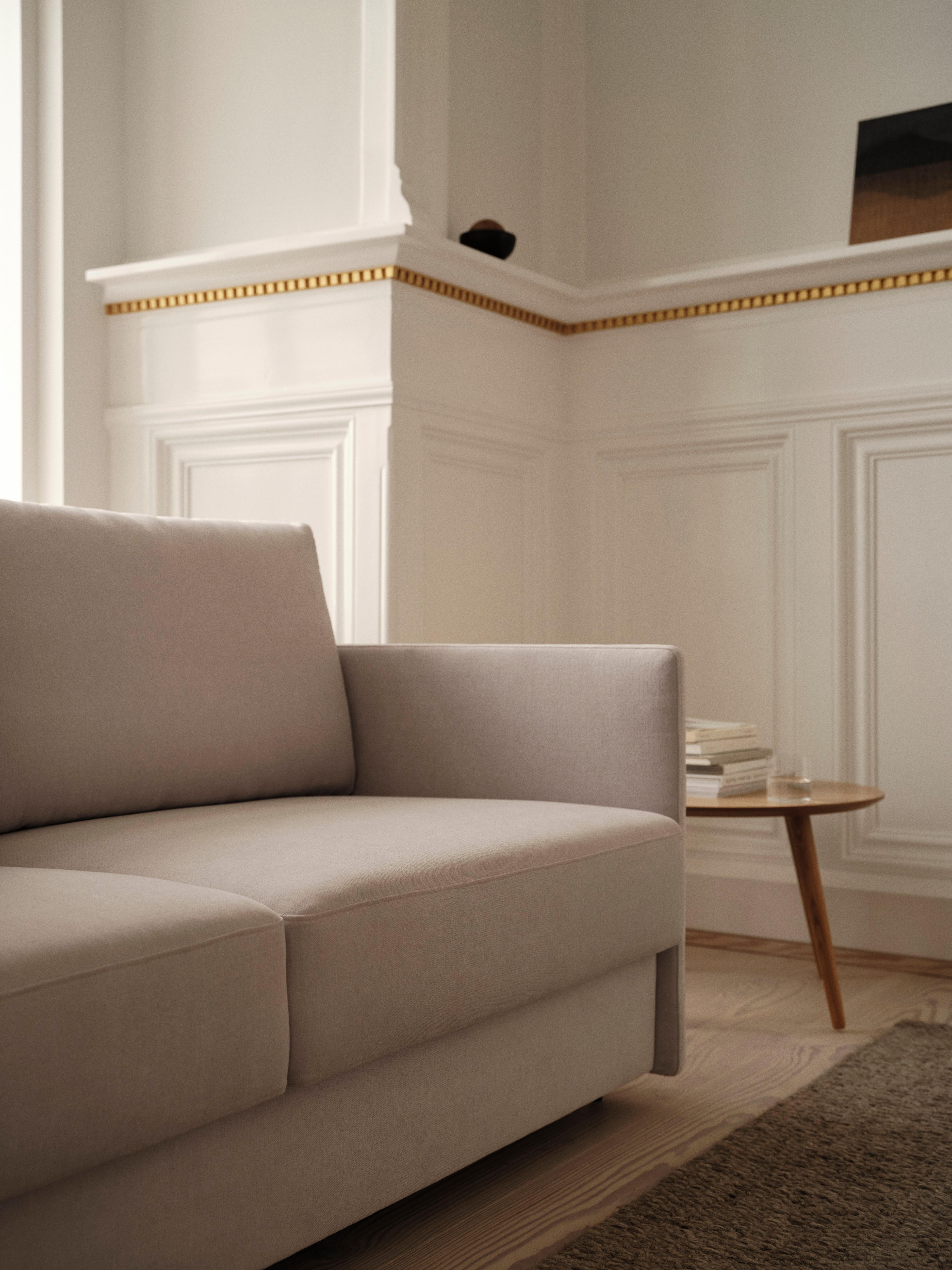 Beige sofa beside a wooden table with stacked books in a room with white panel walls.