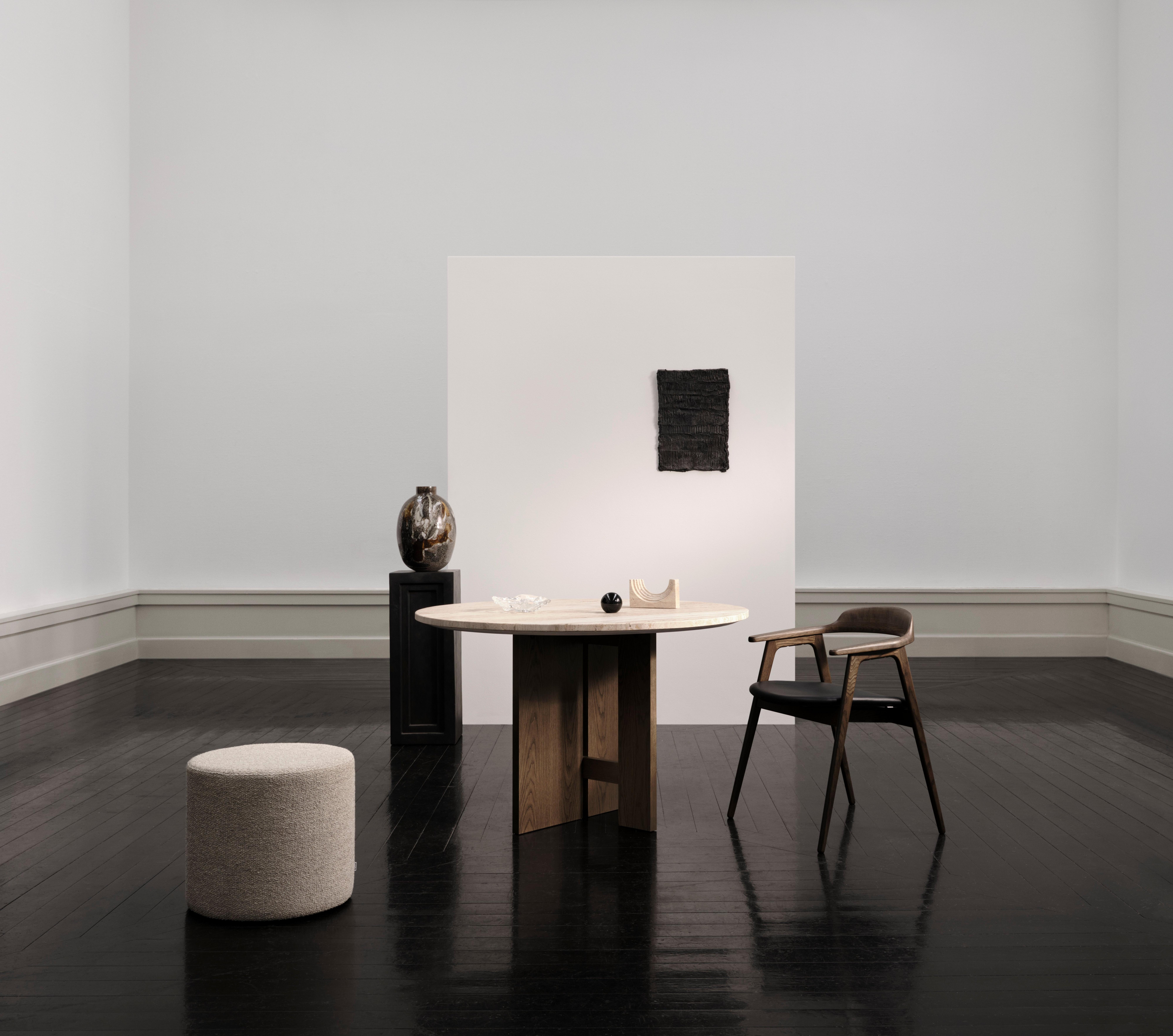 Holztisch mit Deko, Stuhl, Hocker und Vase auf dunklem Boden vor weißer Wand. Kunstwerk hängt an Wand.