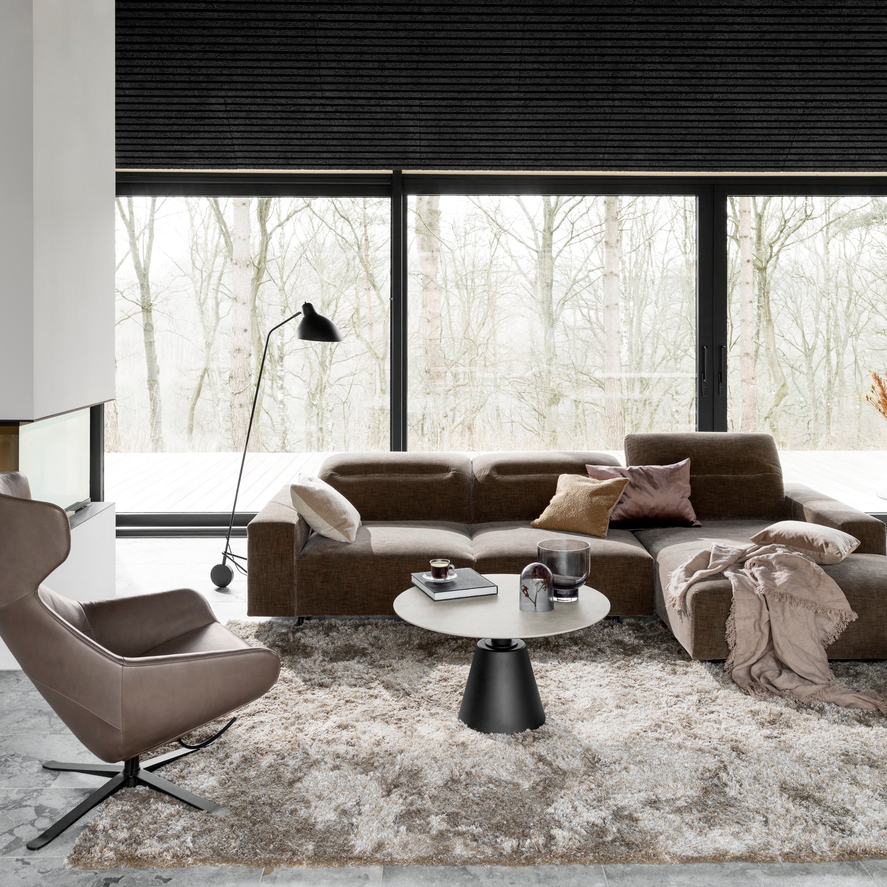 Sala moderna con sofá seccional marrón, alfombra gris y lámpara de pie negra junto a la ventana.
