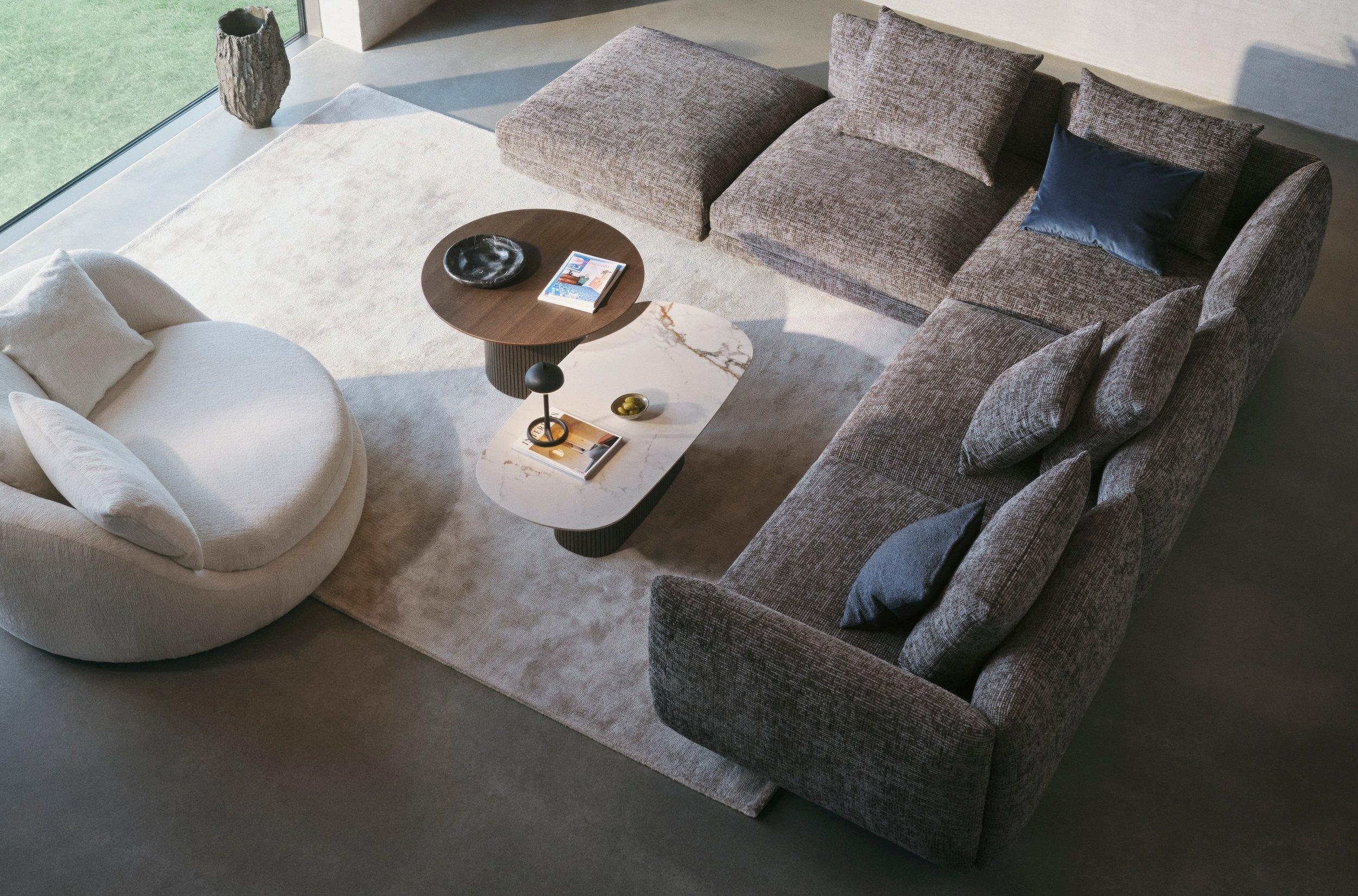 Moderní obývací pokoj s šedou texturovanou modulární pohovkou, kulatým bílým křeslem a stohovatelnými stolky na světlém koberci u velkého okna.