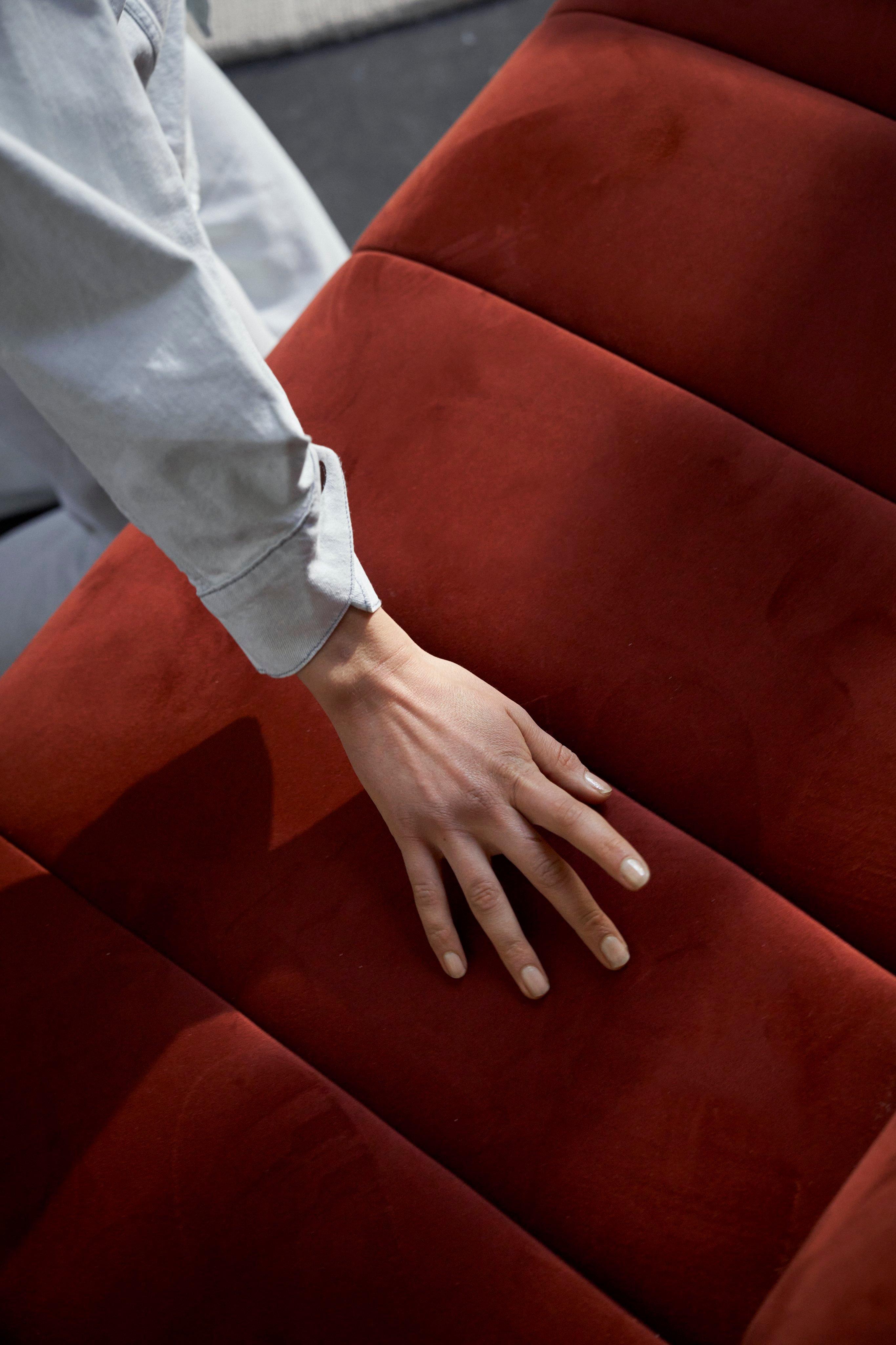 Személy kezével érinti a Bellagio kétszemélyes kanapé piros bársony kárpitozását, érzékeltetve lágy textúráját és modern dizájnját.