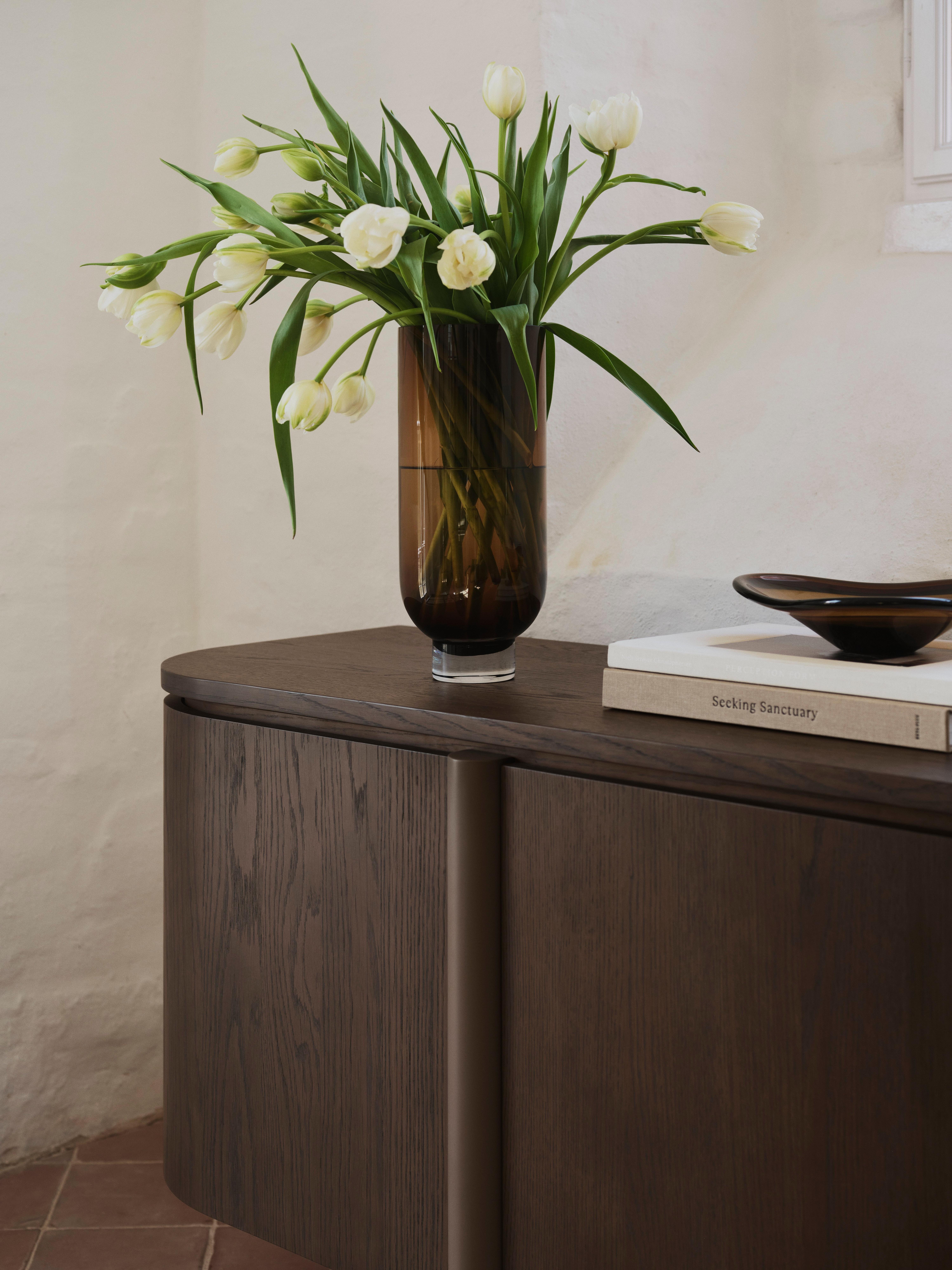 White tulips in a tall brown glass vase on a wooden sideboard beside stacked books.