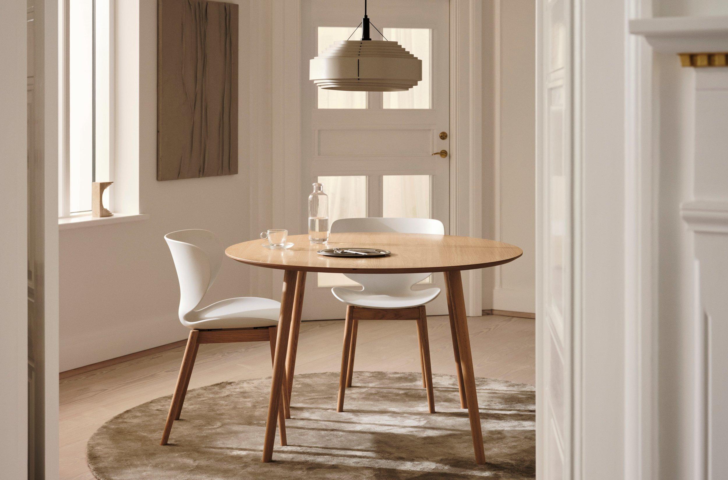 Round wooden table with two white chairs. Pendant light above, glass bottle and cup on table.