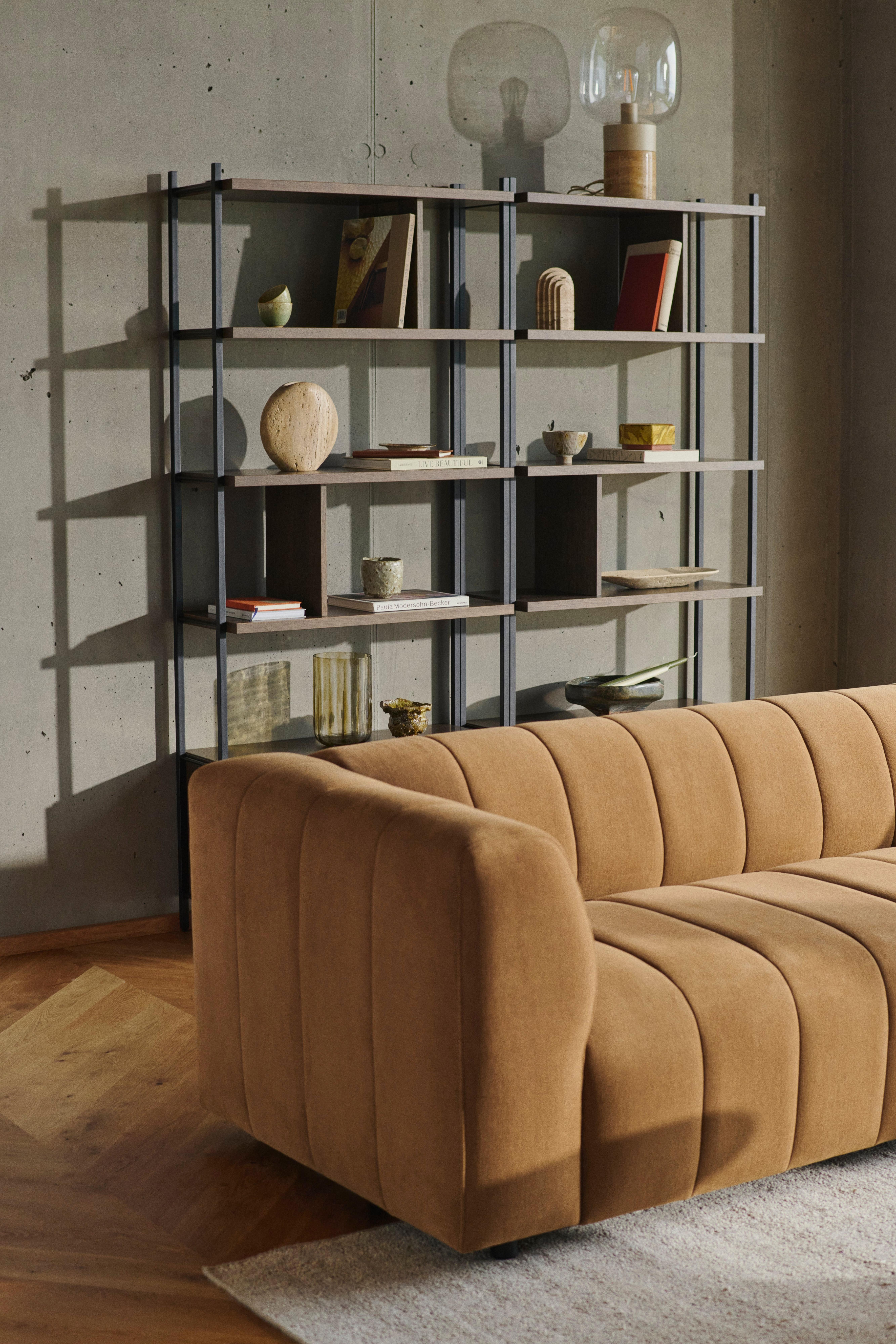 Nahaufnahme eines getufteten Bellagio Sofas neben einem schwarzen Metallregal mit Dekoartikeln, Büchern und einer Glaslampe an einer Betonwand.