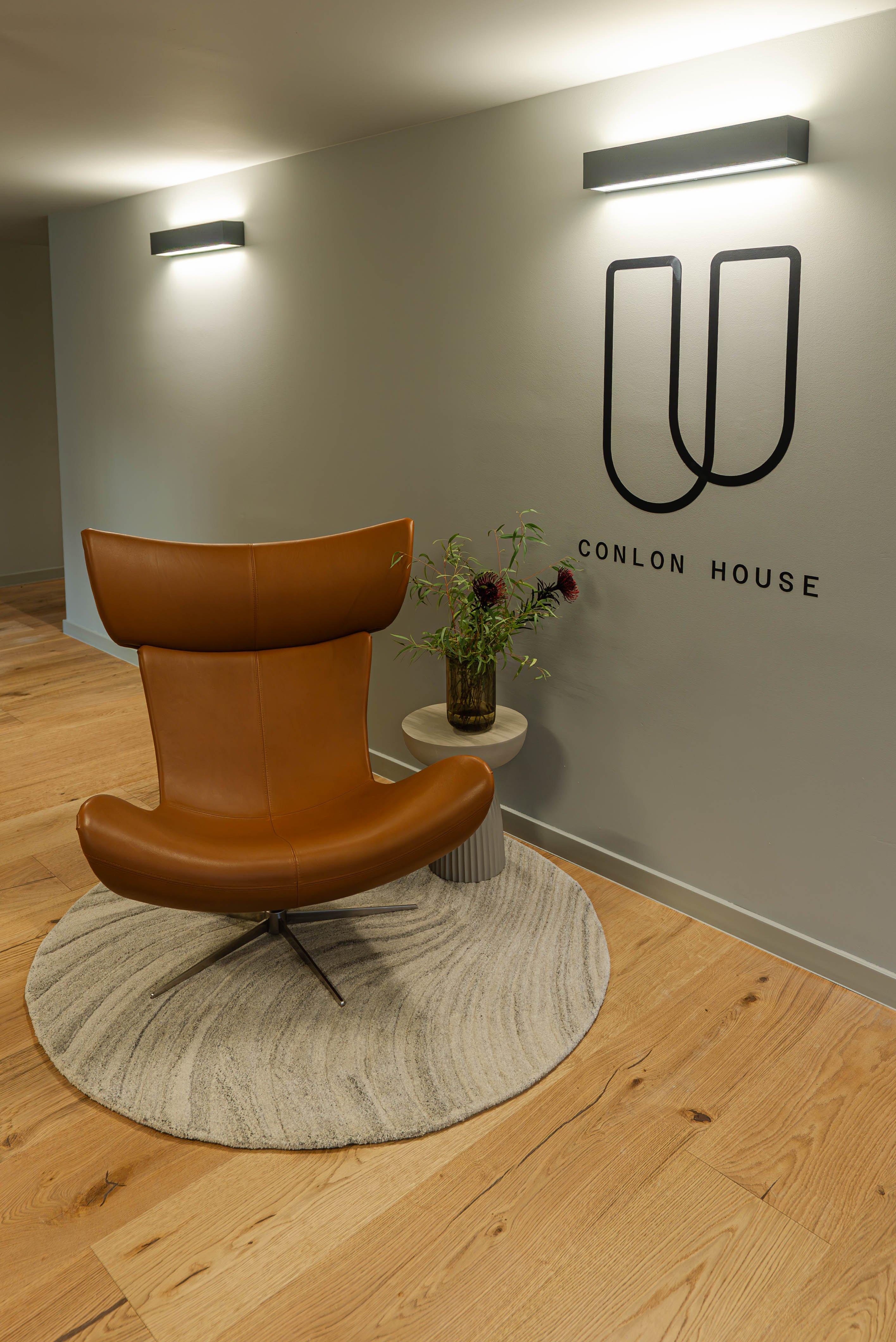 Fauteuil en cuir marron sur tapis rond, près d'une table avec vase de fleurs, mur marqué "Conlon House".