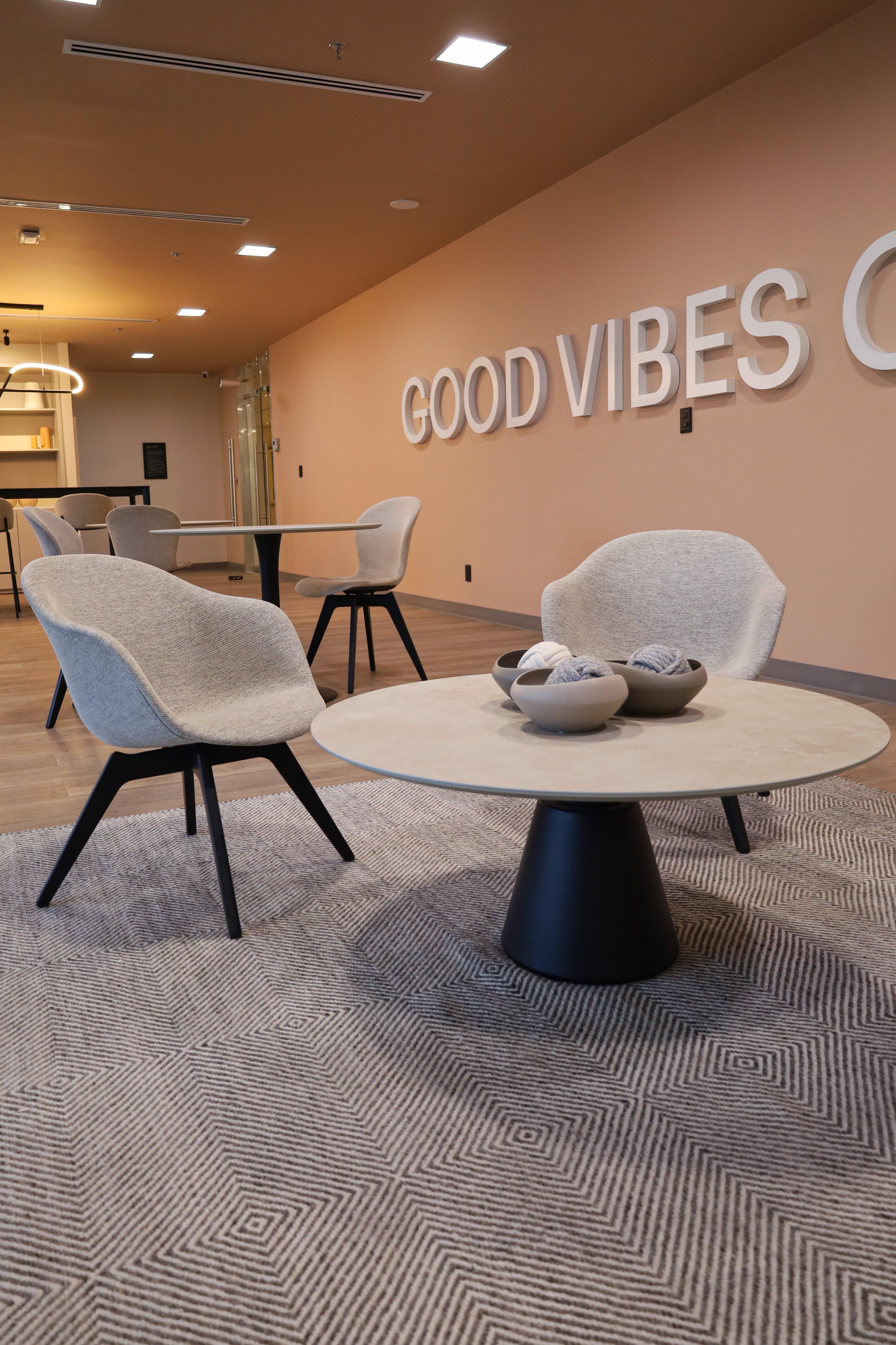 Modern lounge with grey chairs, round tables, and wall text "GOOD VIBES".