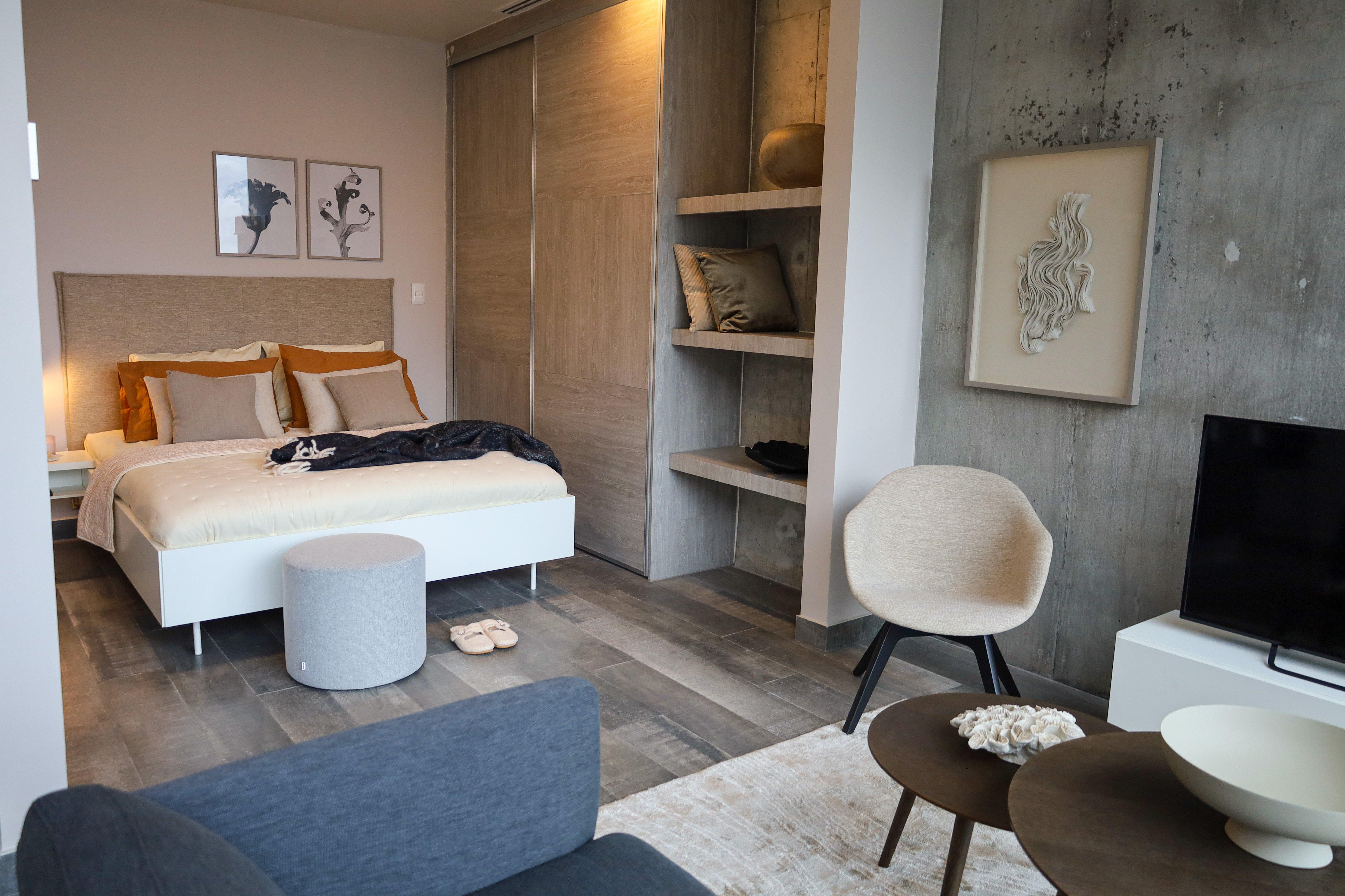 Modern bedroom with a double bed, armchair, TV, and artwork on walls. Shelves hold cushions.