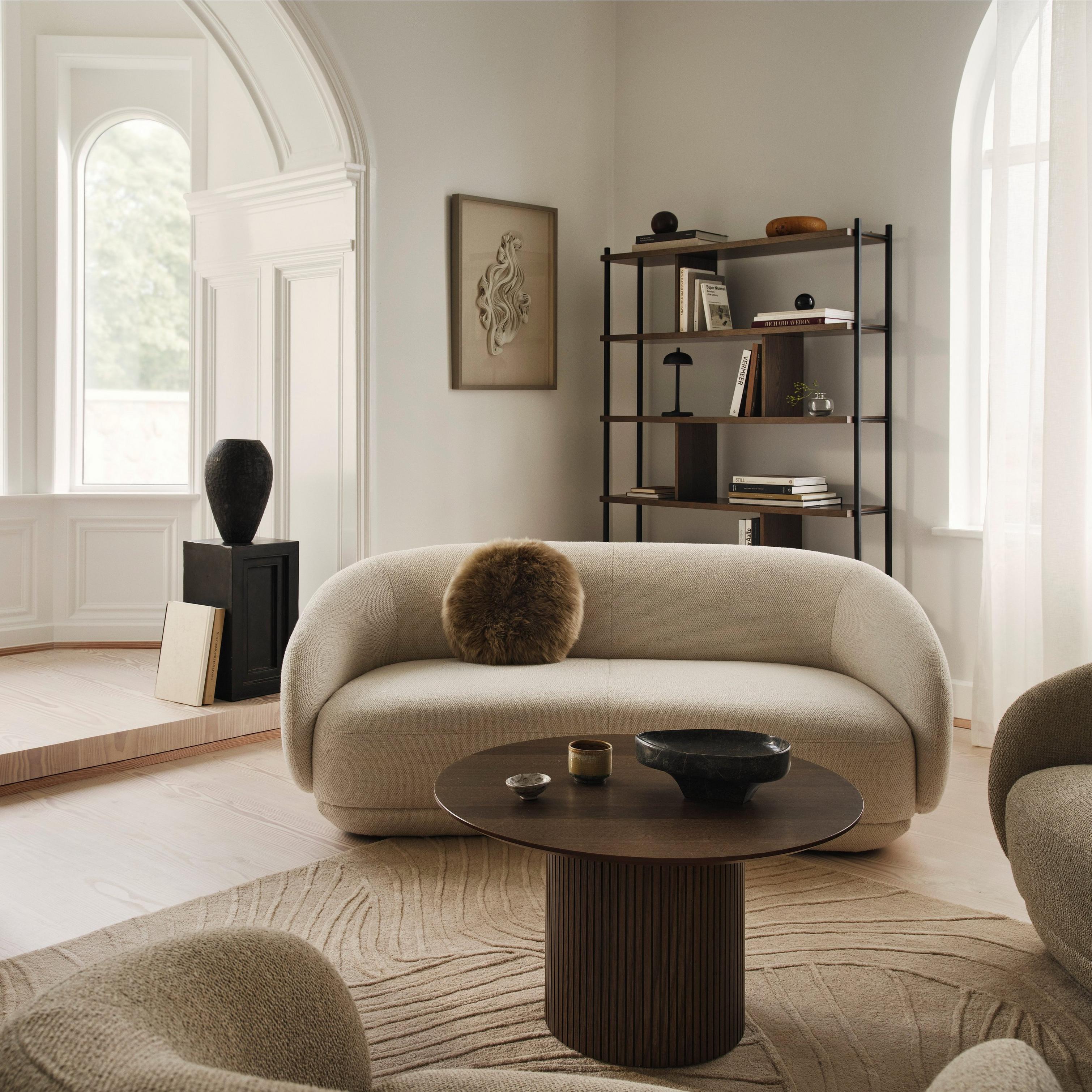 Modern living room with a beige sofa, round table, and bookshelf. Large windows provide natural light.