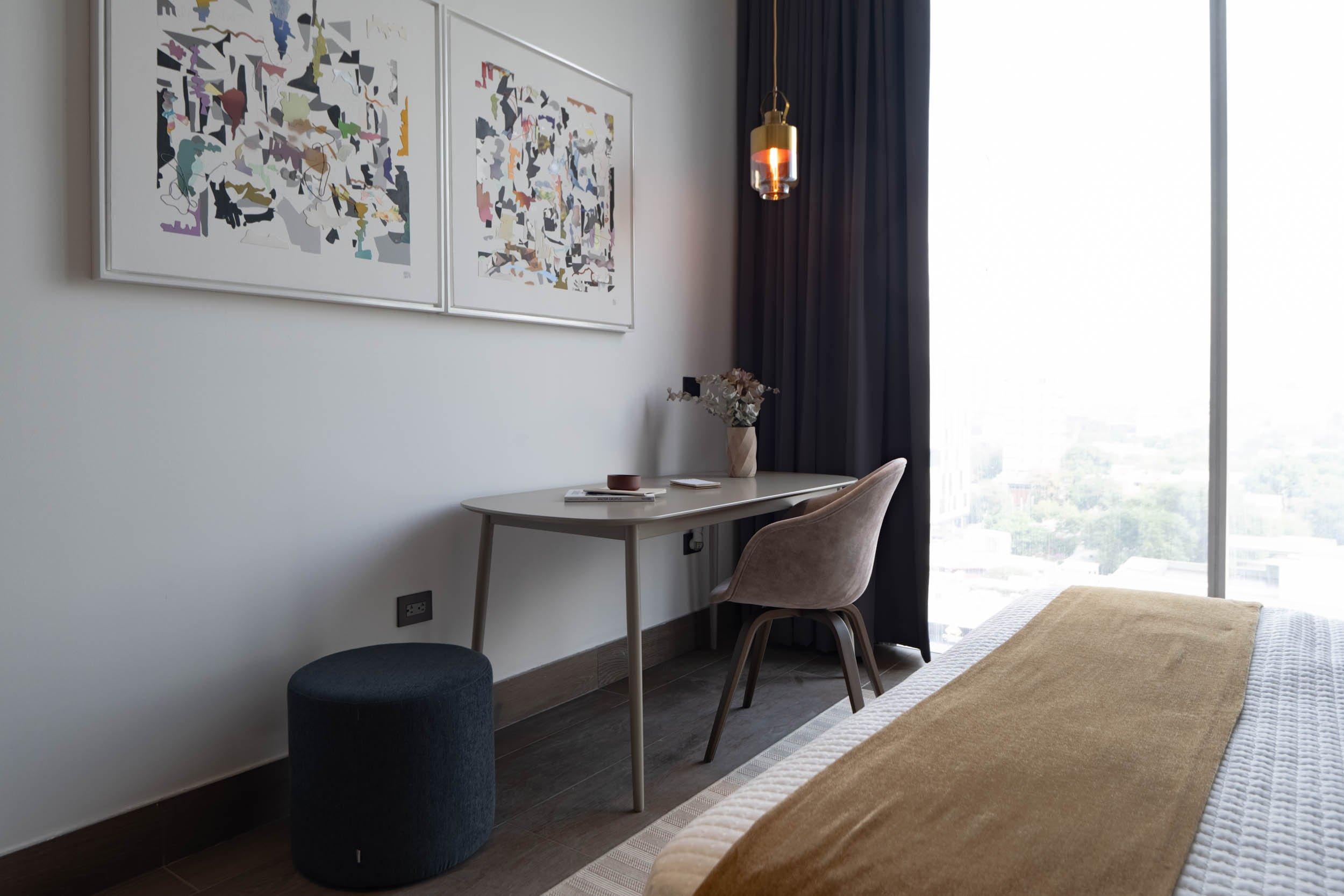 Bureau avec chaise près d’une fenêtre, lampe suspendue et cadres abstraits au mur.