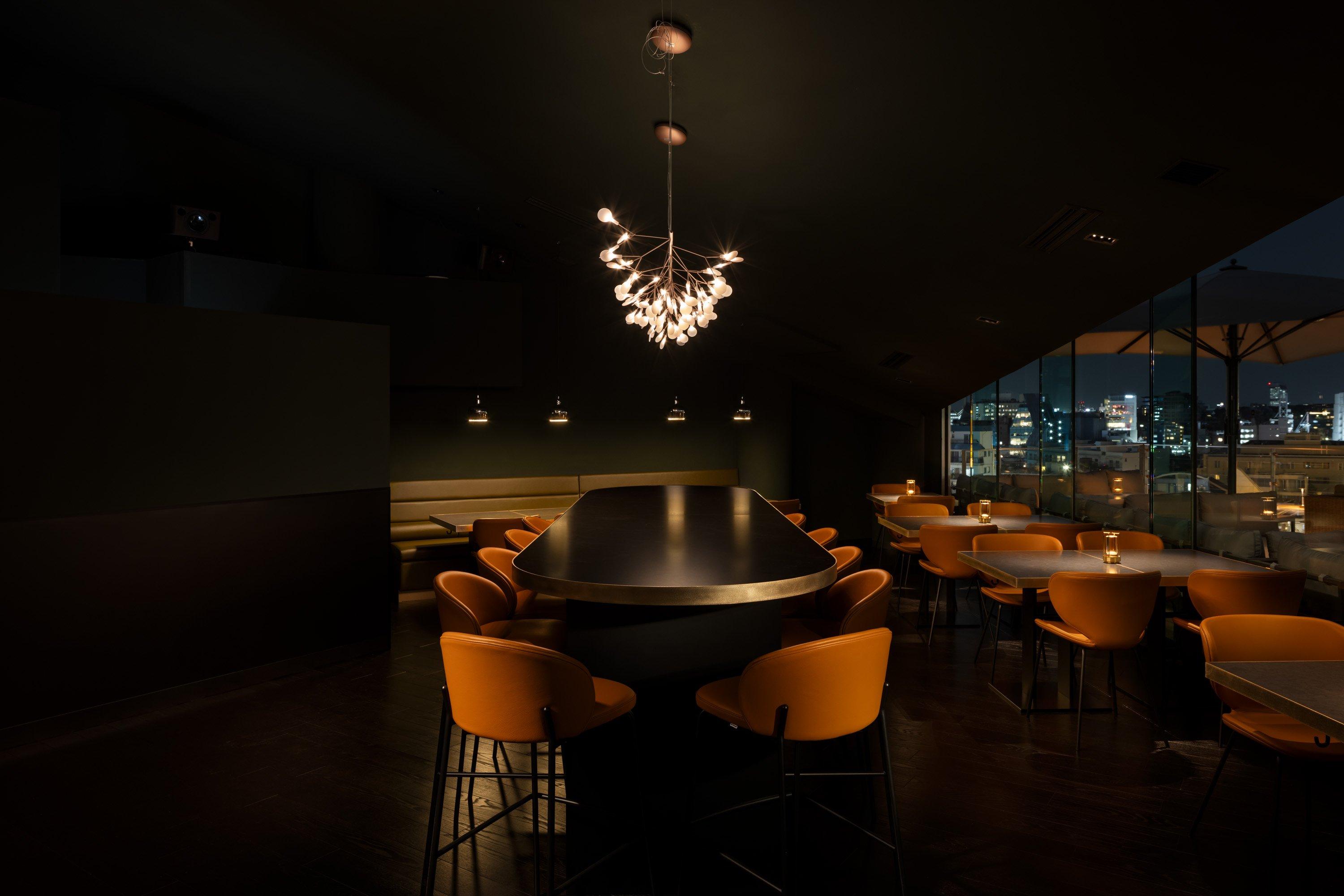 Sala de restaurante elegante con iluminación tenue, sillas naranjas y vista nocturna de la ciudad.