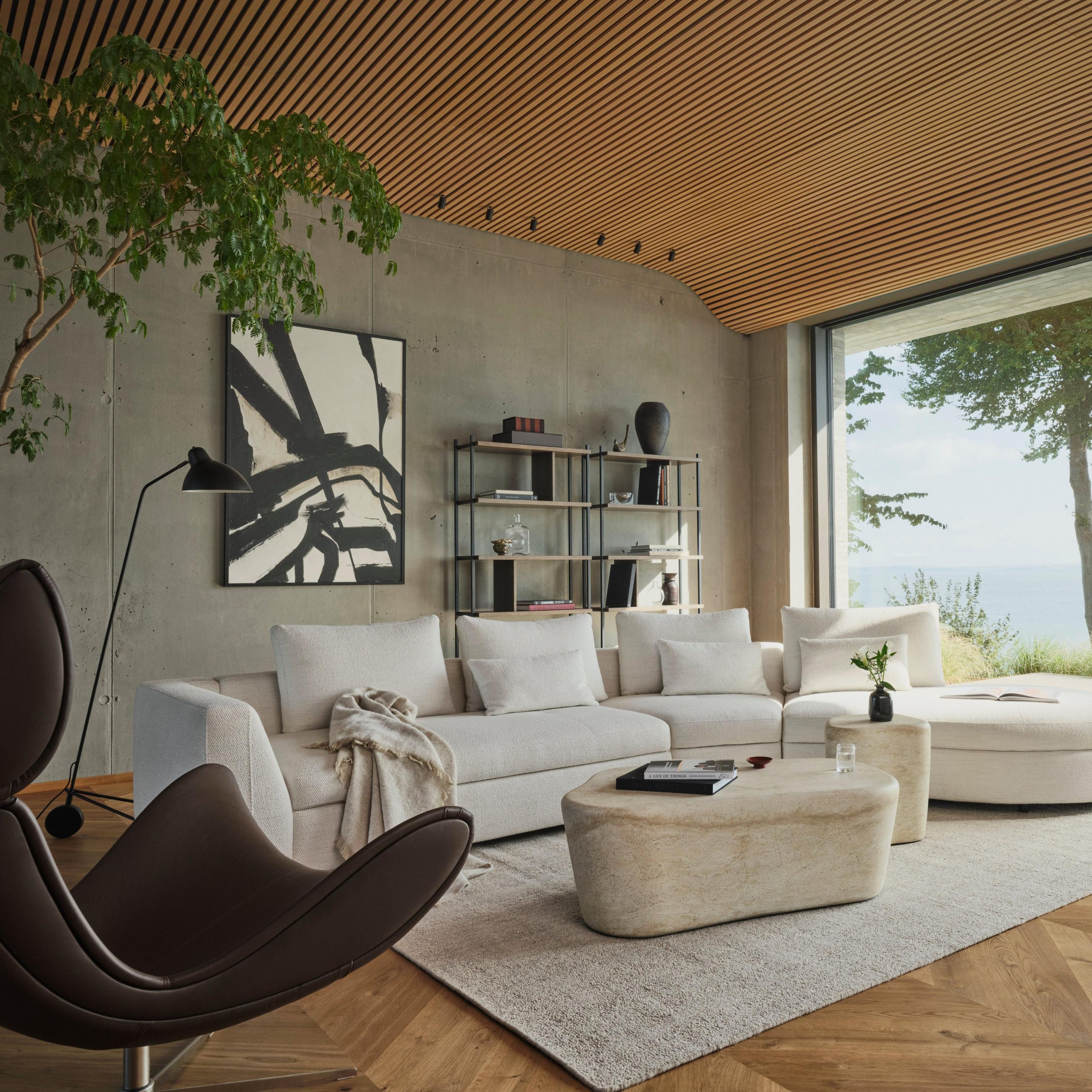 Modern living room with beige sofa, abstract art, large window view of trees and sea.